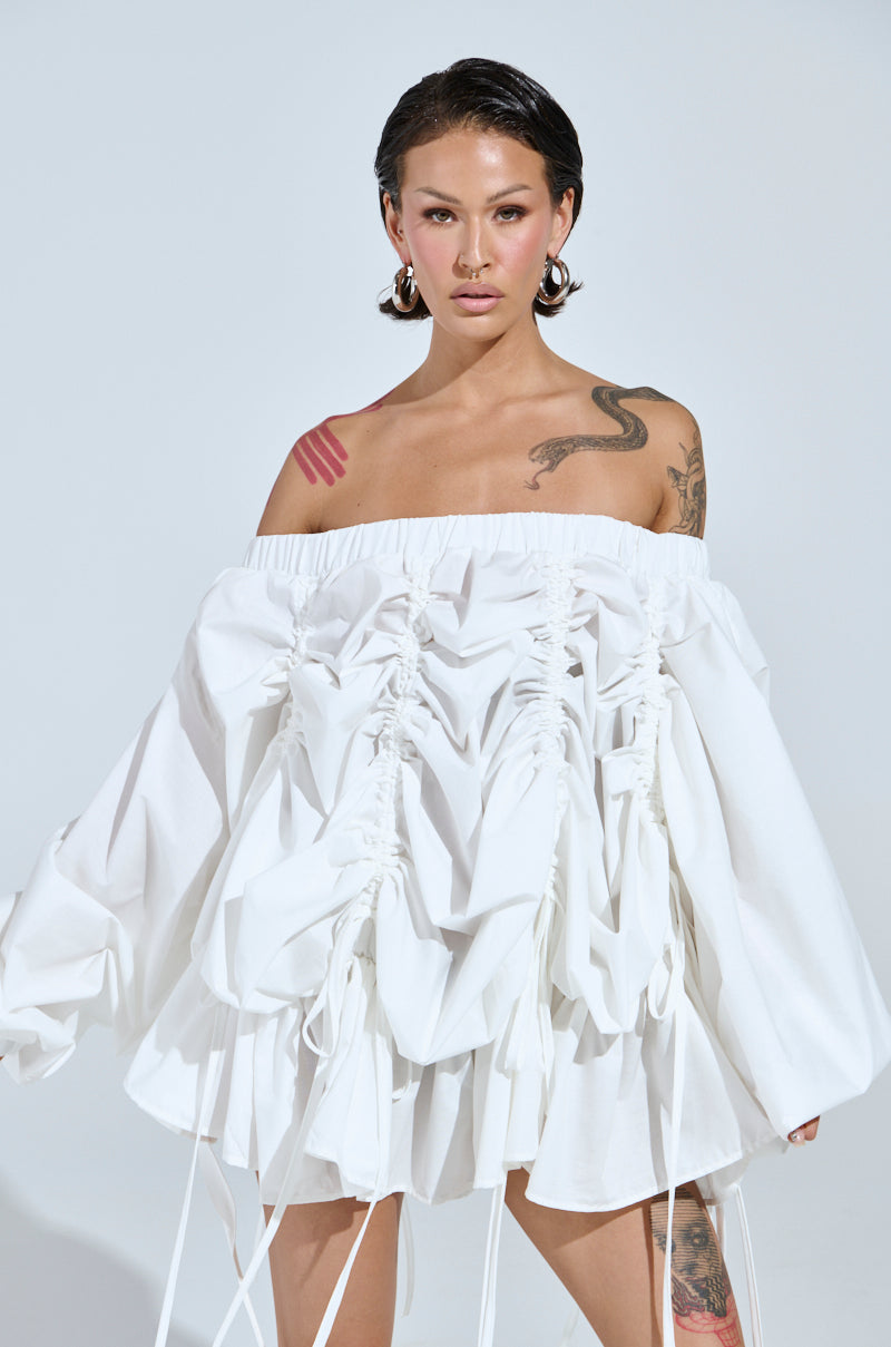 A person stands against a light background wearing the "Free As Can Be" white mini dress with ruching and off-the-shoulder design. They have a snake tattoo on one arm, large hoop earrings, short hair styled back, and a pink abstract tattoo on their shoulder.