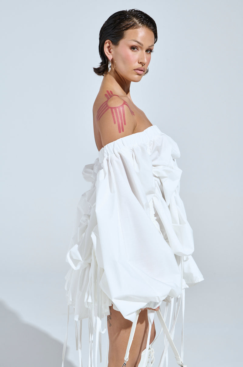 A woman models the FREE AS CAN BE MINI DRESS IN WHITE with a slicked-back hairstyle and hoop earrings. She stands in profile, showcasing a pink abstract design on her arm against a plain light gray background.