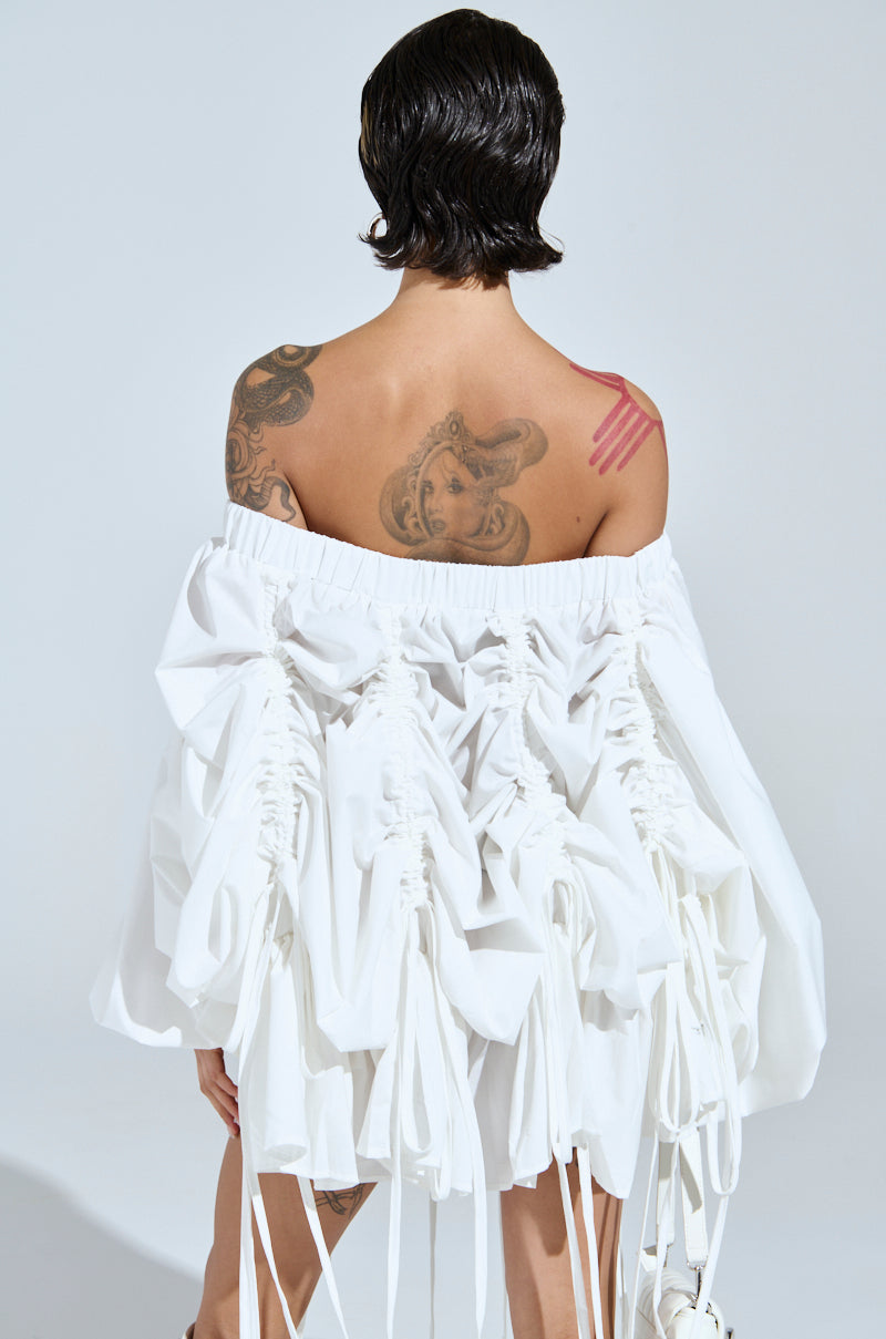 A person with short dark hair faces away from the camera, wearing the FREE AS CAN BE MINI DRESS IN WHITE—a white off-the-shoulder mini dress with gathered ruffles and ties. Tattoos decorate their back as they hold a white object in one hand.