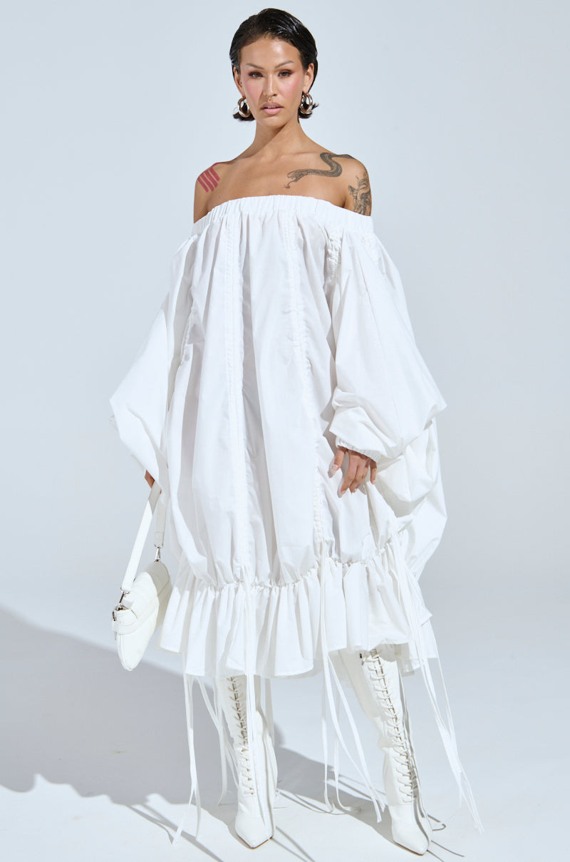 Wearing the FREE AS CAN BE MINI DRESS IN WHITE, an off-the-shoulder piece with long sleeves and a ruffled hem, they pair it with large hoop earrings, white knee-high lace-up boots, and a matching handbag. Visible tattoos on the left shoulder enhance the ensemble's elegant allure.