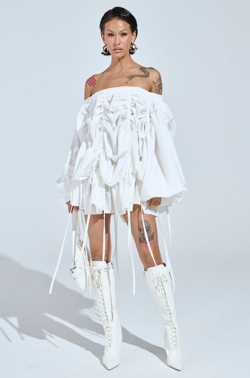 Wearing the FREE AS CAN BE MINI DRESS IN WHITE and tall white lace-up boots, with tattoos on arms and thighs, they hold a sleek white handbag against a minimalist white backdrop.