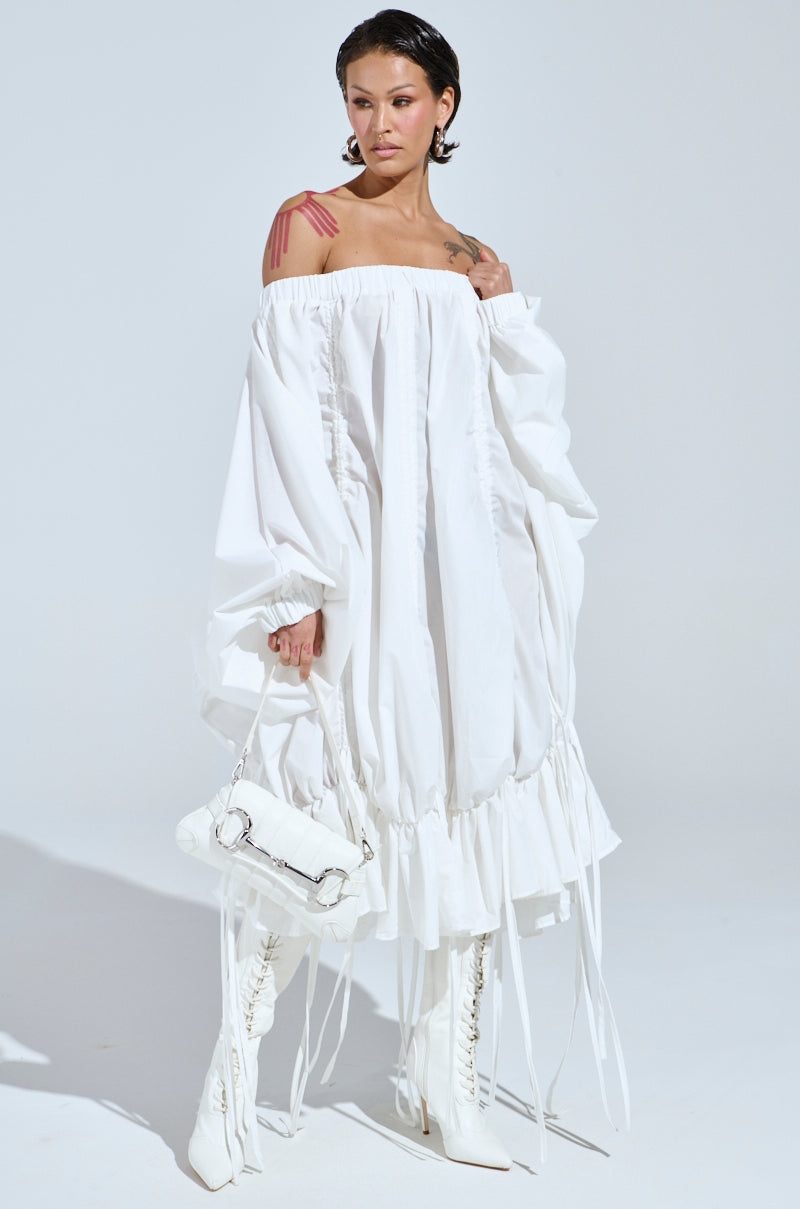 Wearing the "FREE AS CAN BE MINI DRESS IN WHITE," a person confidently poses in an off-the-shoulder, loose-fitting, white mini dress with long sleeves. They hold a white handbag and wear knee-high white boots, showing their tattooed arm against a light gray background with slicked-back hair.