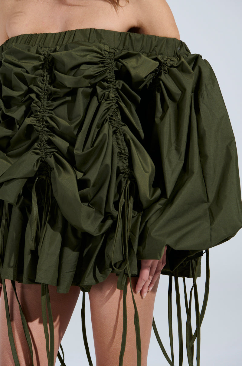 The individual is wearing the "FREE AS CAN BE MINI DRESS IN OLIVE," an off-the-shoulder mini dress in dark green with dramatic ruching, gathers, and oversized sleeves. The voluminous fabric features long ties at the edges, revealing their arms below.