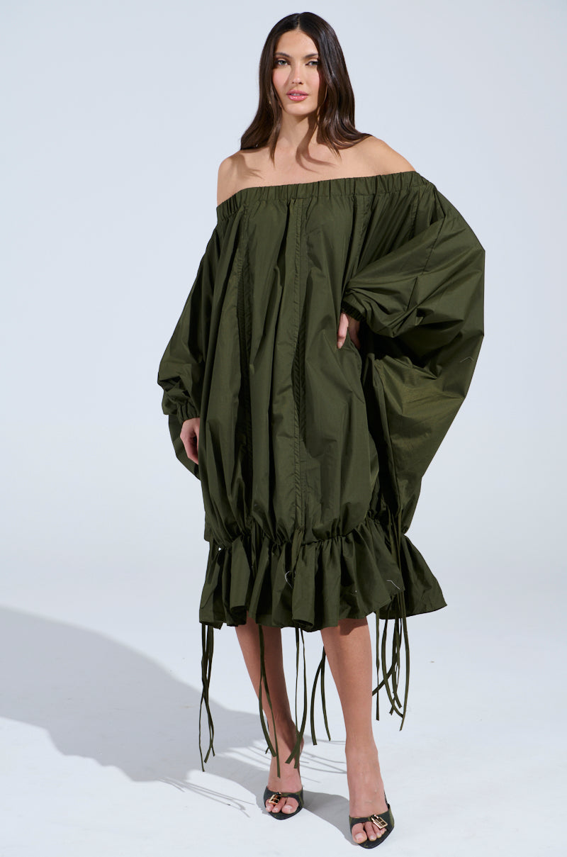 A woman poses in the FREE AS CAN BE MINI DRESS IN OLIVE, featuring off-the-shoulder styling, a ruffled hem, and ruched details. She completes the look with black heels against a light gray background.