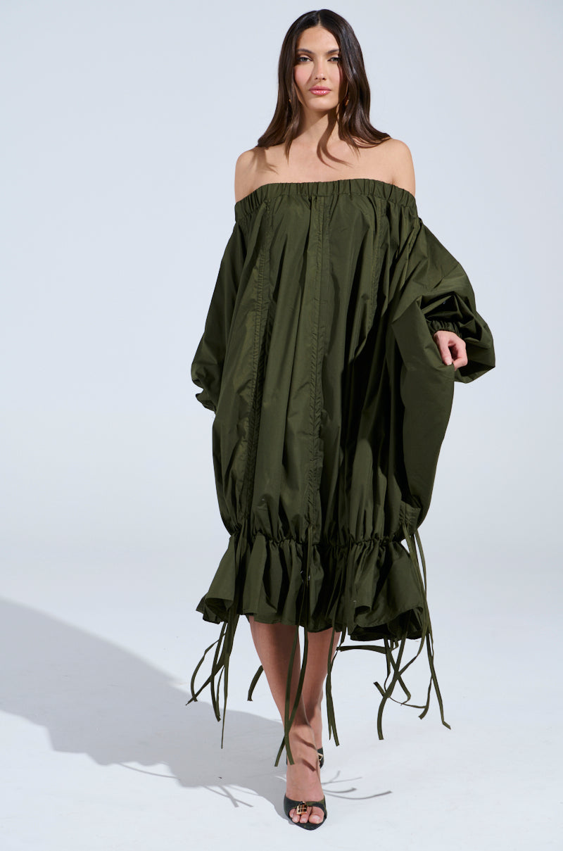 A woman with long brown hair wears the FREE AS CAN BE Mini Dress in olive, featuring an off-the-shoulder design and adjustable drawstring. She walks confidently on a plain white background, her minimal makeup enhancing her style, perfectly paired with black high-heeled sandals.