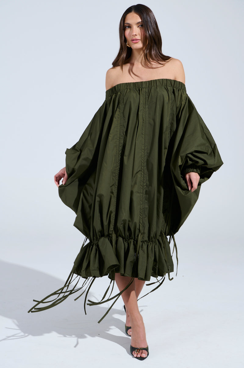 A woman poses in the FREE AS CAN BE MINI DRESS IN OLIVE, featuring a voluminous off-the-shoulder design with ruffled hem and loose sleeves. This elegant dress pairs perfectly with her black heels as she stands against a plain white background.