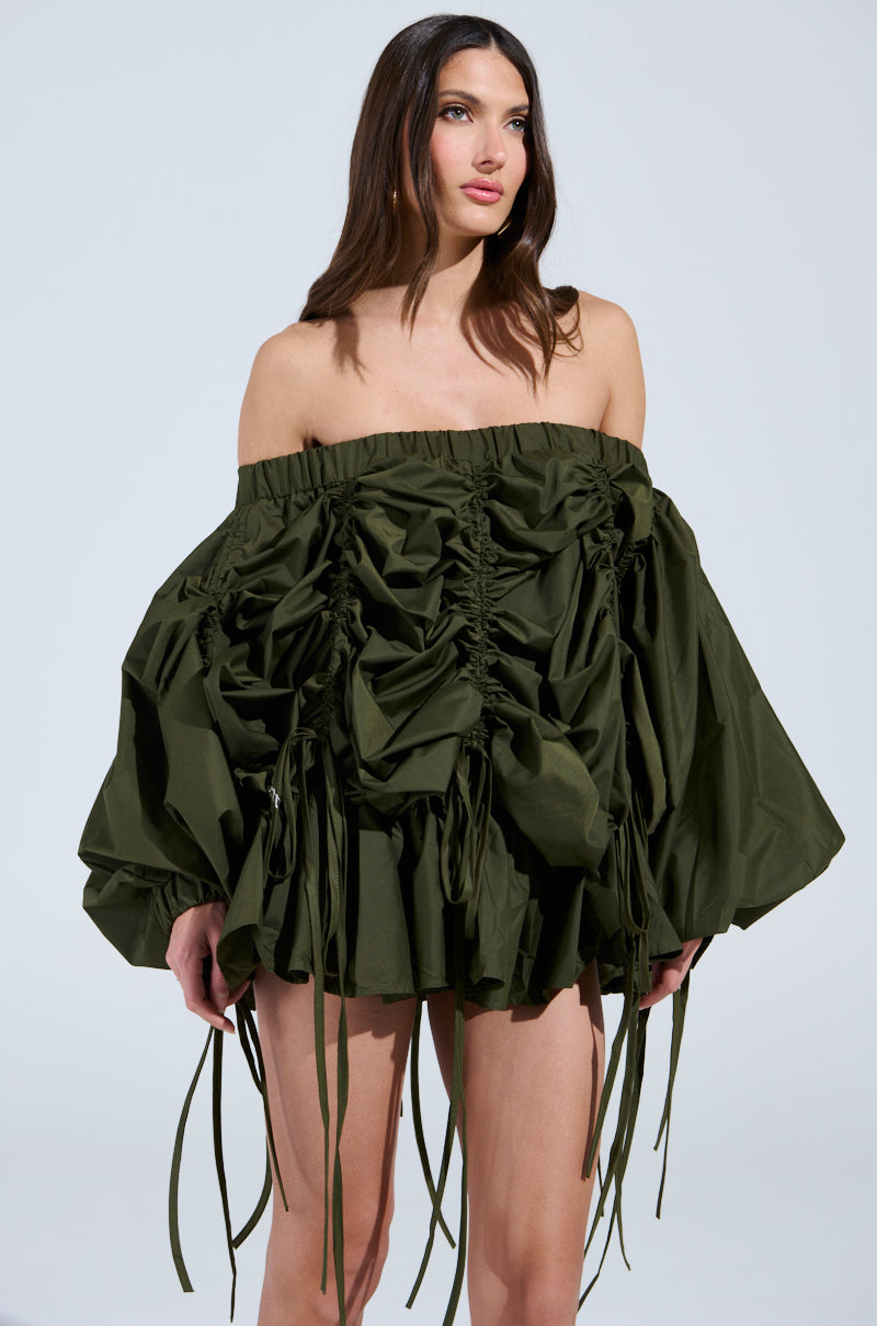 A woman in a ruched, off-the-shoulder olive mini dress with dramatic hanging ribbons and long voluminous sleeves stands with long brown hair and a neutral expression. The dress is the "Free As Can Be Mini Dress in Olive.