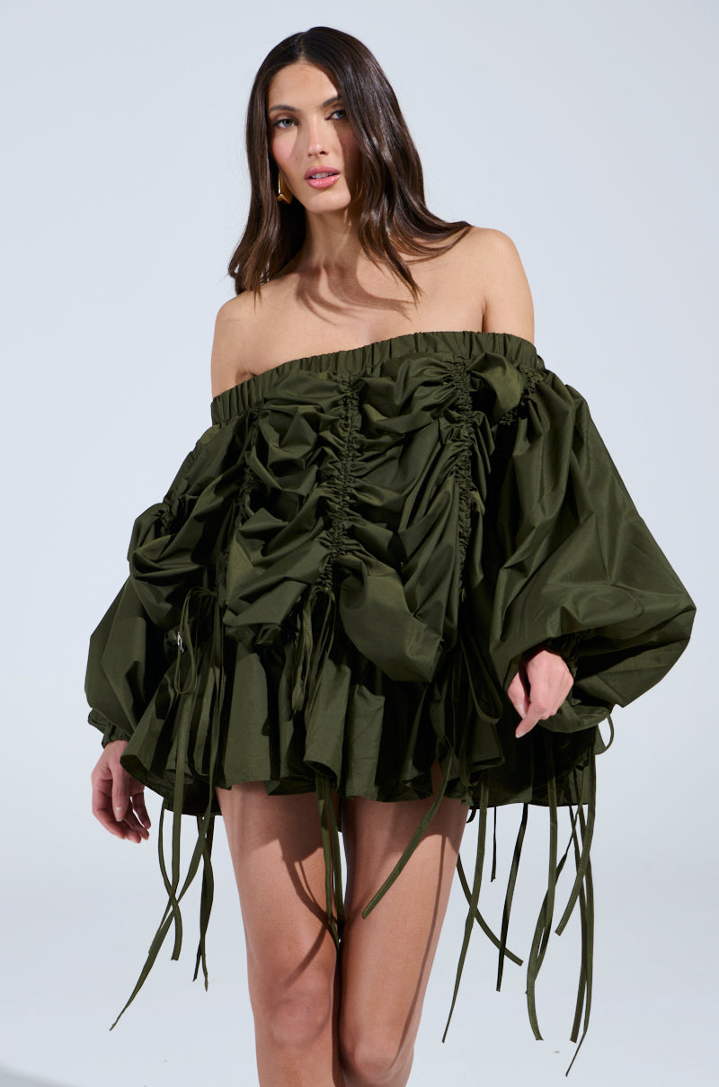 A woman poses in the FREE AS CAN BE MINI DRESS IN OLIVE, featuring an off-the-shoulder design with ruched fabric, voluminous sleeves, and hanging ribbons. Her long, dark hair stands out against the plain background.