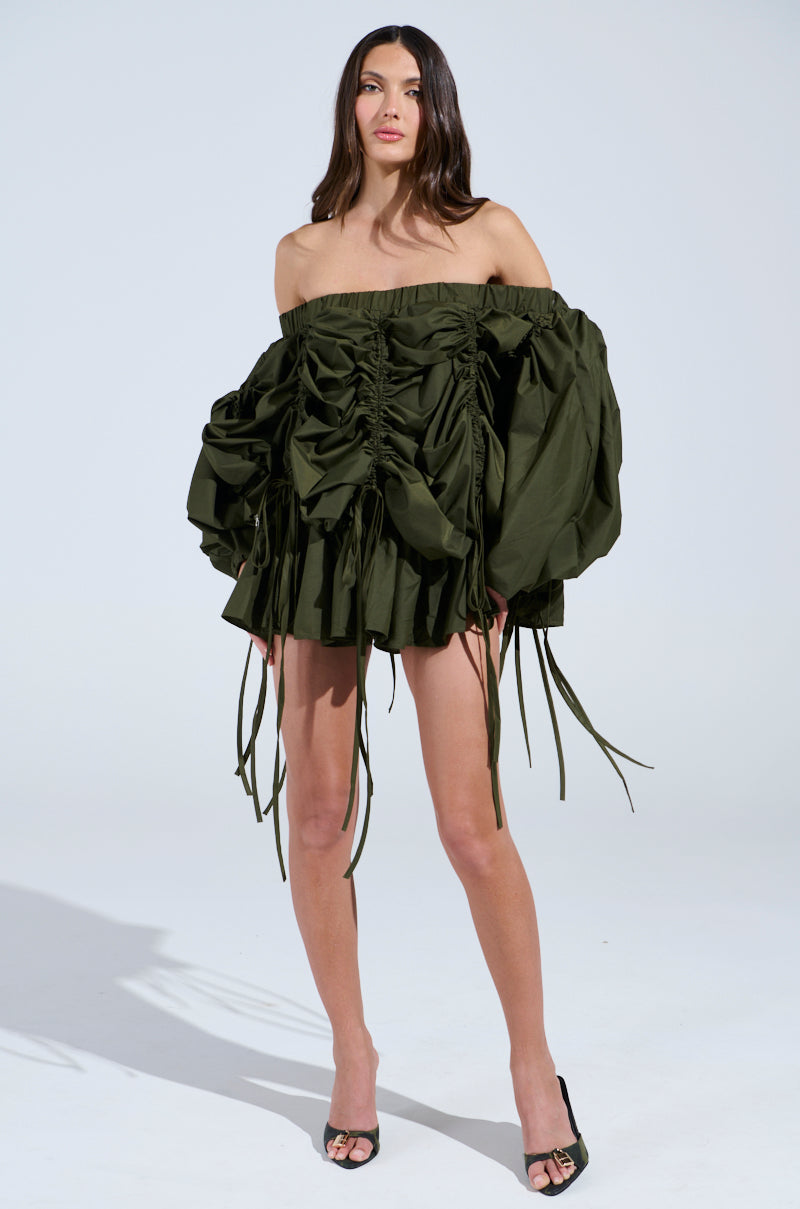 A person confidently wears the FREE AS CAN BE MINI DRESS IN OLIVE, an off-the-shoulder, dark green mini dress with ruched design and long ribbons. They're paired with black open-toe heels against a light gray backdrop.