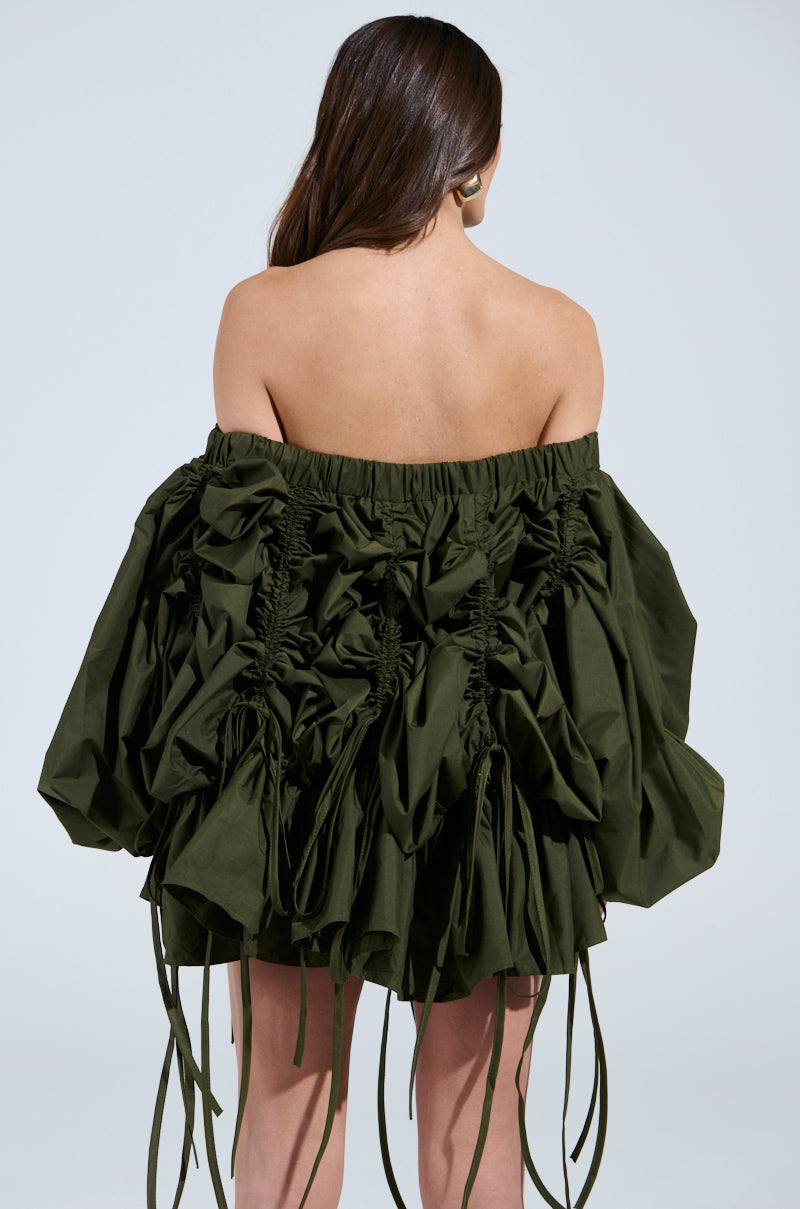 A person with long dark hair, facing away from the camera, wears the FREE AS CAN BE MINI DRESS IN OLIVE. The ruched design adds texture, and elongated ties drape elegantly from the bottom, perfectly complemented by a soft light gray background.