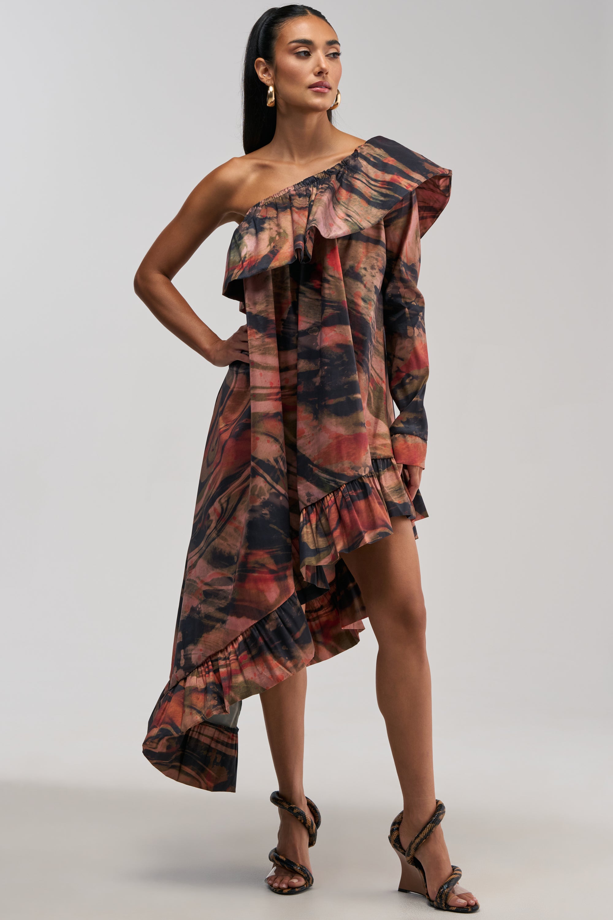 Model wearing the MOOD SWING PRINTED BUBBLE MINI DRESS, featuring an asymmetrical hem, off-shoulder neckline, and ruffled mini dress detailing in an abstract red, brown, and black print, styled with strappy heels against a plain background.