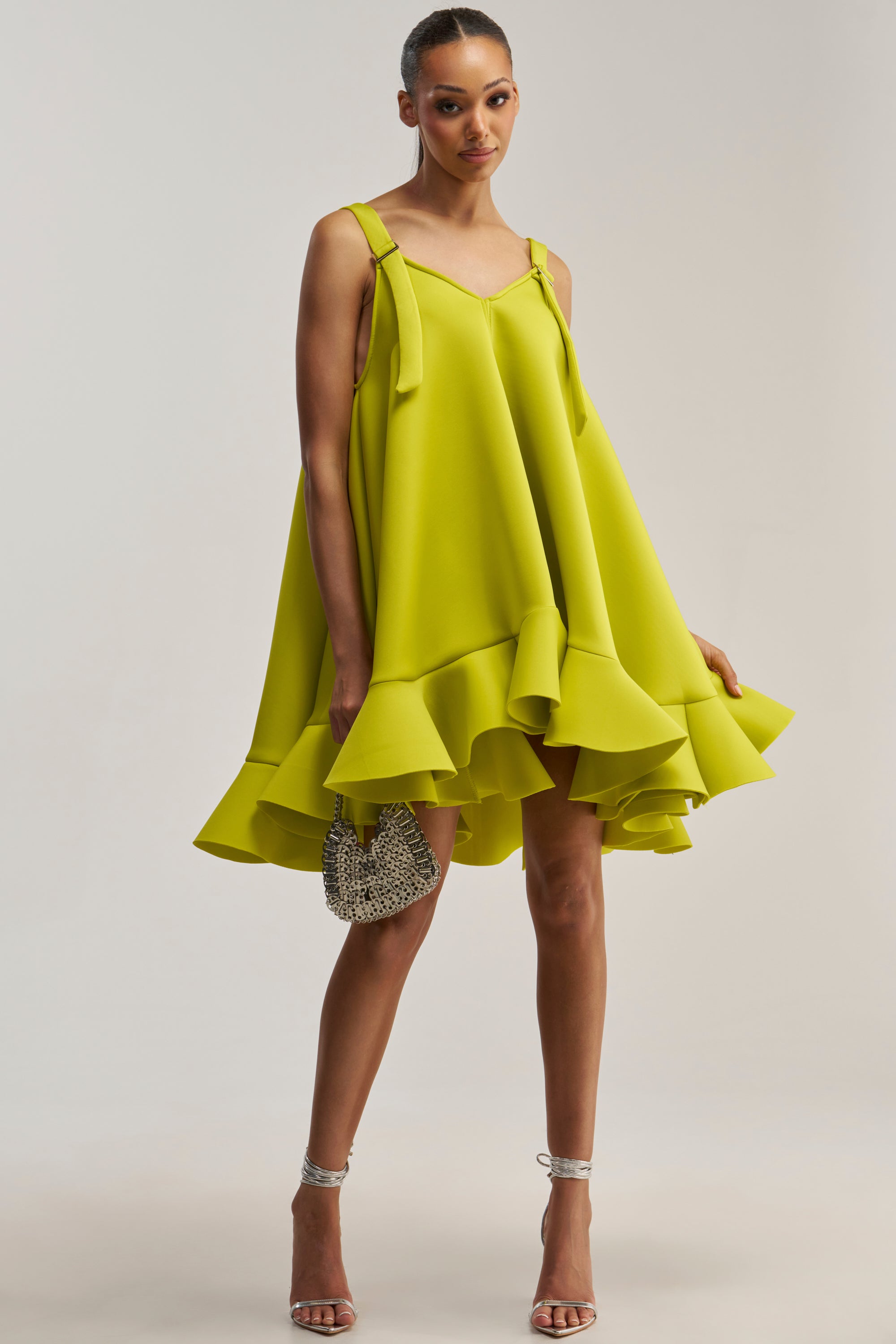 A woman wearing the IN TOO DEEP SCUBA MINI DRESS IN CHARTREUSE, featuring a loose fit and ruffled hemline, holds a small beaded purse and stands in silver heels against a plain background.