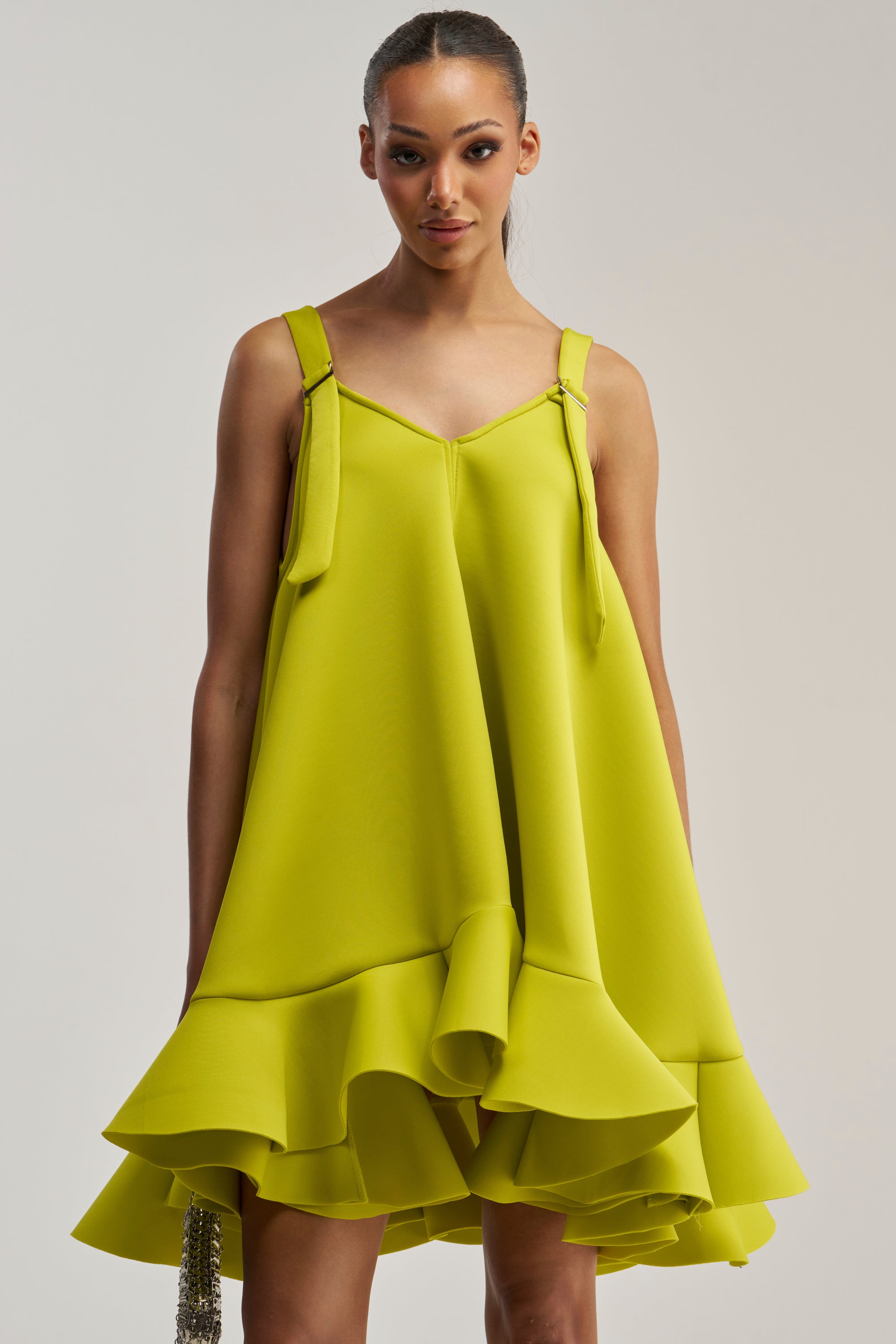 A woman wears the IN TOO DEEP Scuba Mini Dress in Chartreuse, a sleeveless dress with shoulder straps and a ruffled hemline, standing against a light background.
