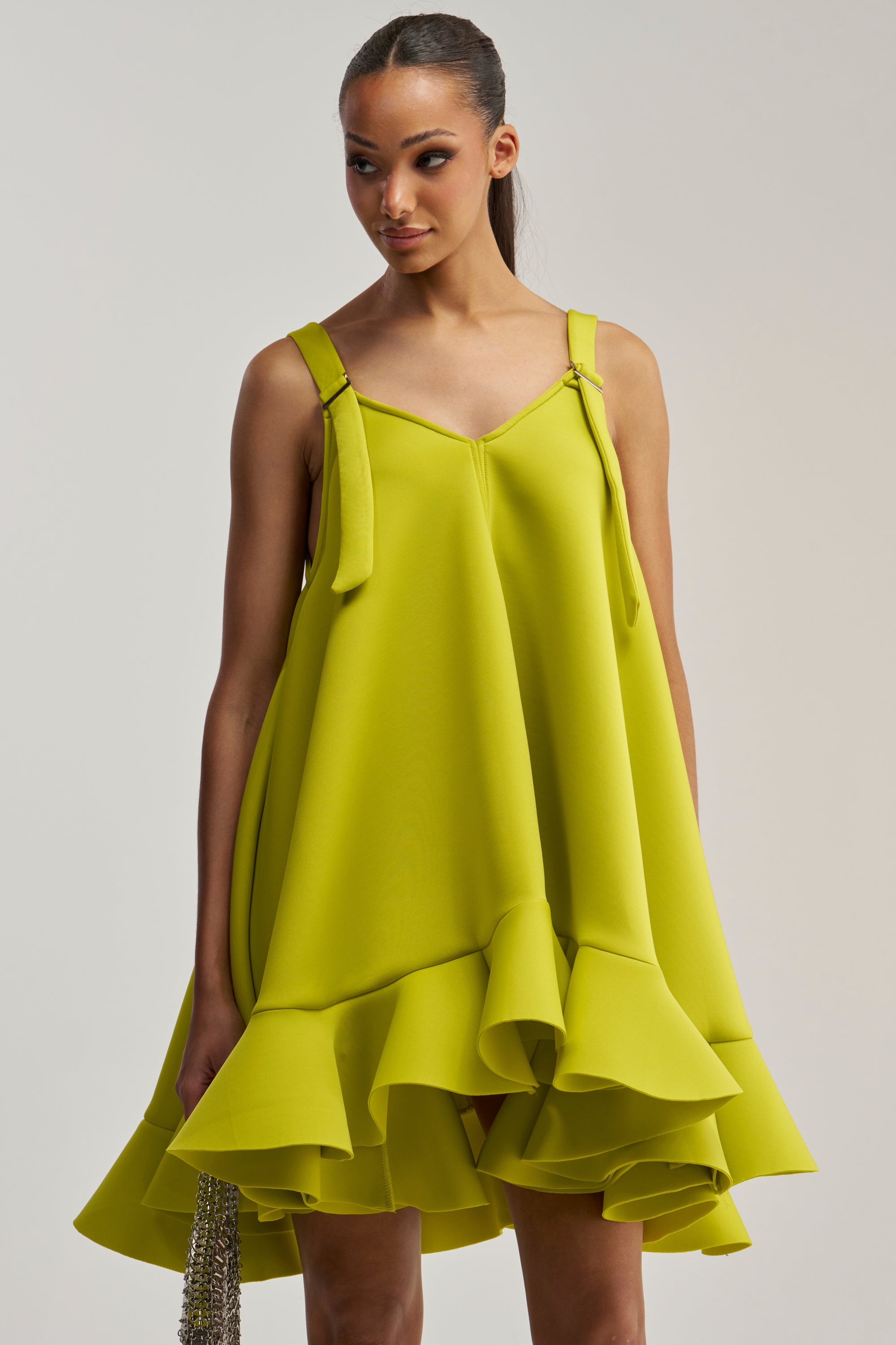 A woman wearing the IN TOO DEEP SCUBA MINI DRESS IN CHARTREUSE, featuring a ruffled hemline and playful bow straps, stands against a plain background while holding a beaded handbag.
