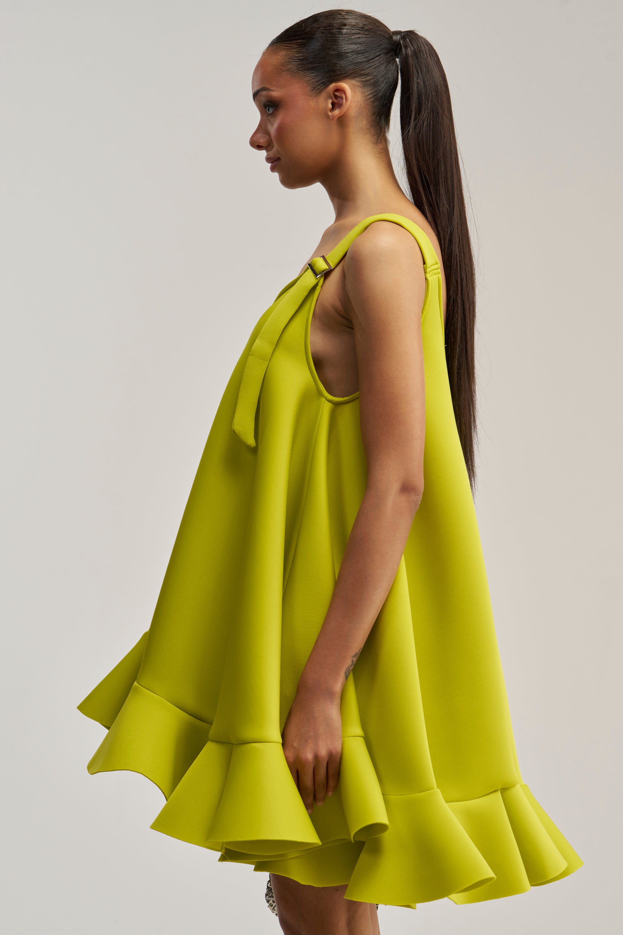 A woman with a long ponytail stands in profile against a plain background, wearing the IN TOO DEEP SCUBA MINI DRESS IN CHARTREUSE—a loose, sleeveless mini dress featuring a striking ruffled hemline.