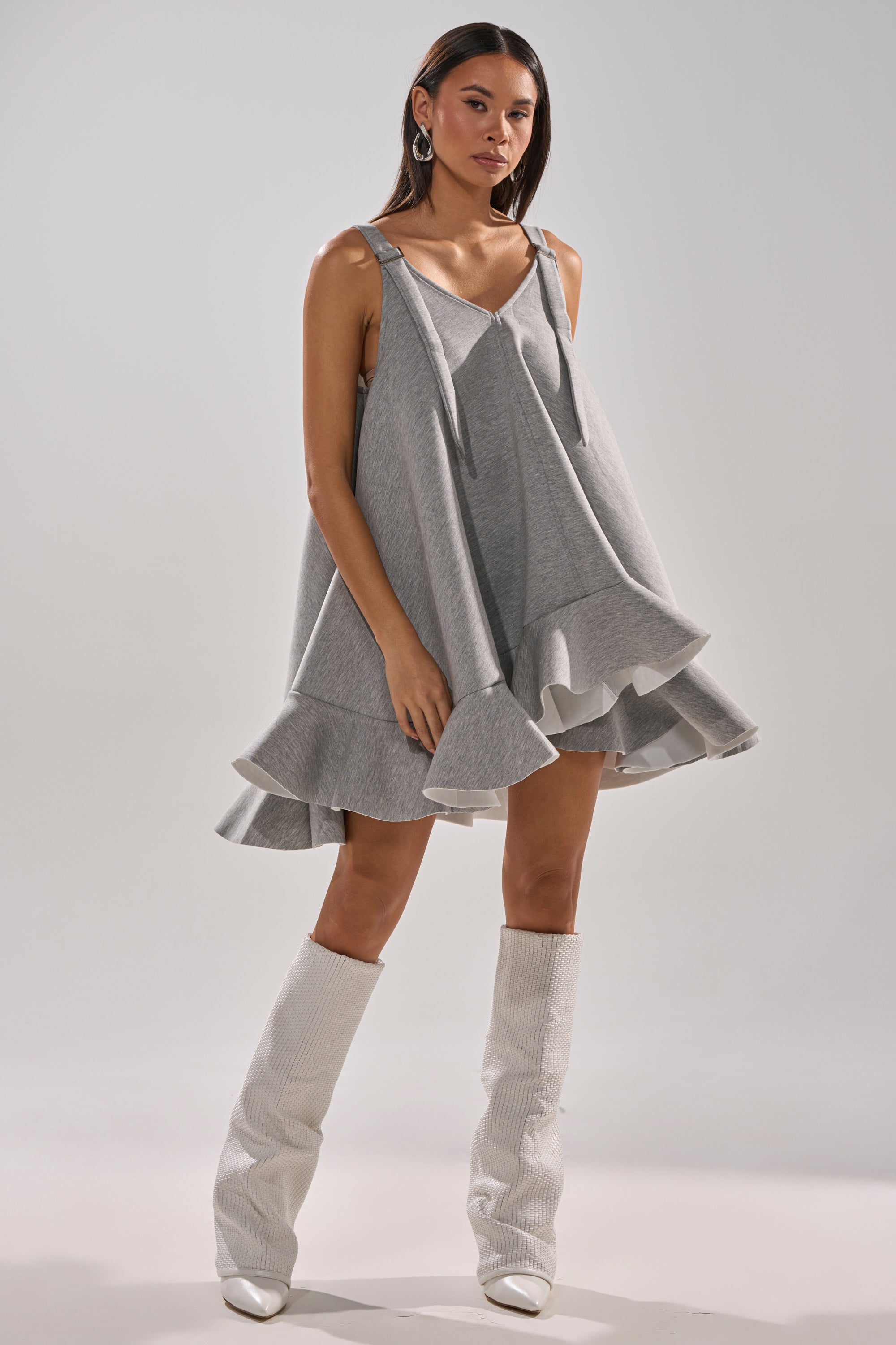A woman poses against a plain light background in the IN TOO DEEP SCUBA MINI DRESS, a loose gray sleeveless mini with a ruffled hem, styled with white knee-high textured boots.