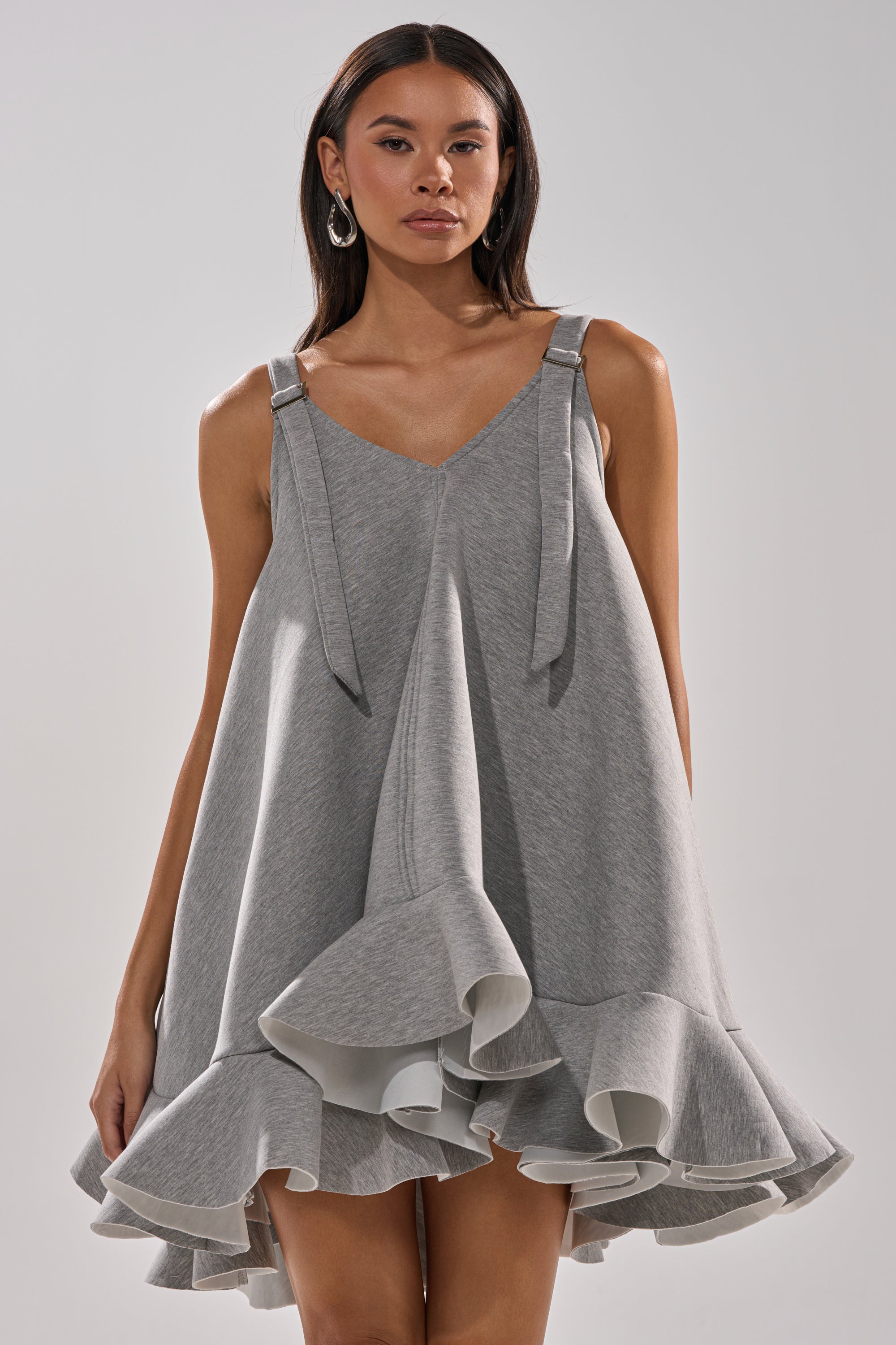 A woman models the IN TOO DEEP SCUBA MINI DRESS, a sleeveless light gray mini dress featuring wide straps and a flared, ruffled hem, while facing forward against a plain light background.