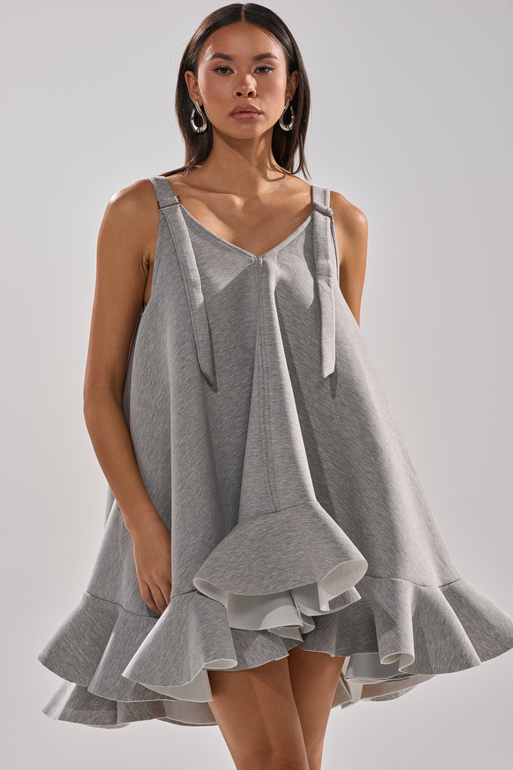 A woman with straight dark hair and hoop earrings poses against a plain background, wearing the IN TOO DEEP SCUBA MINI DRESS—a sleeveless, light grey mini dress featuring an oversized fit, ruffled hem, and bow-detail straps.