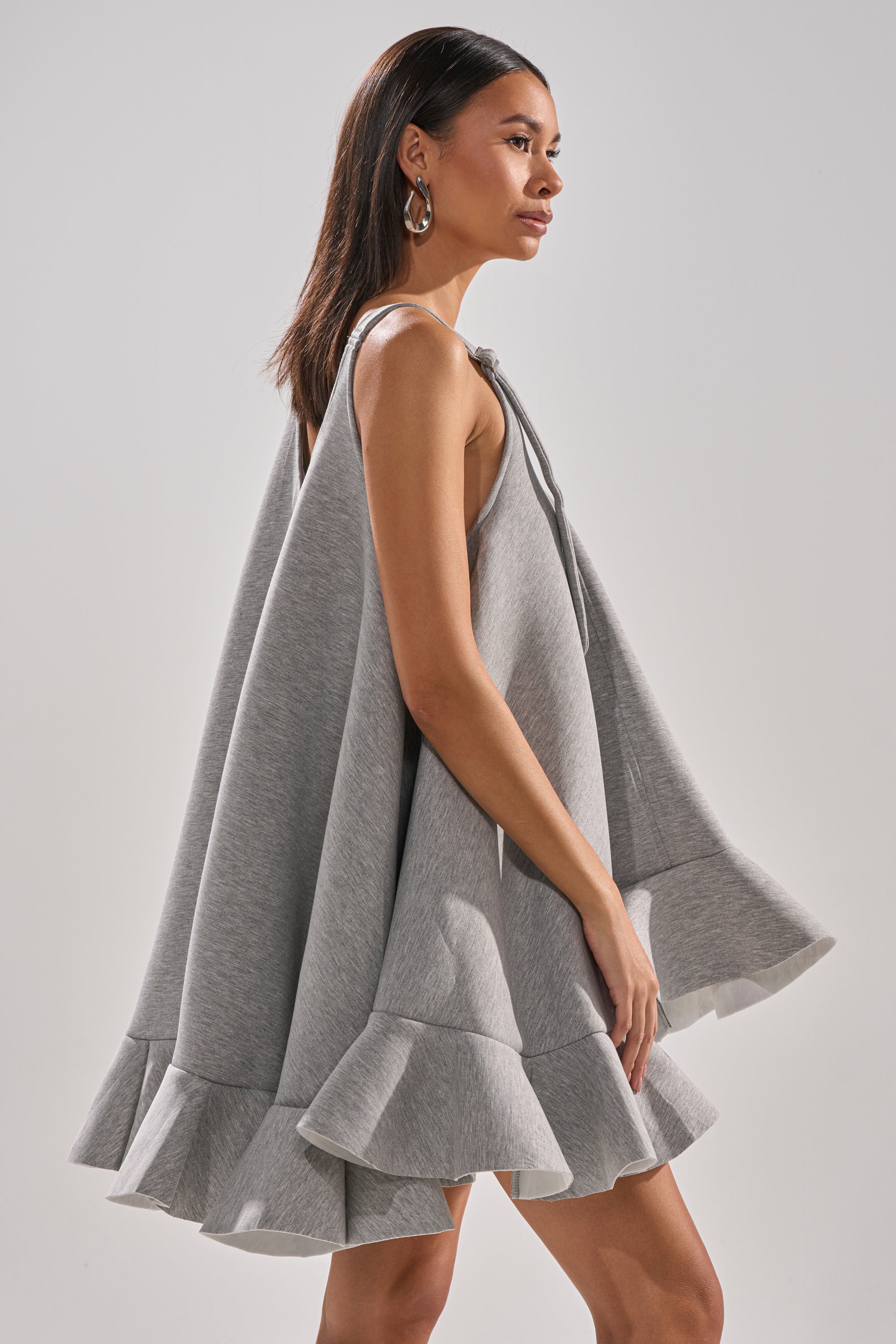 A woman in profile wears the IN TOO DEEP SCUBA MINI DRESS, a sleeveless grey piece with an oversized fit and ruffled hem. She has straight dark hair, hoop earrings, and stands against a plain light background.