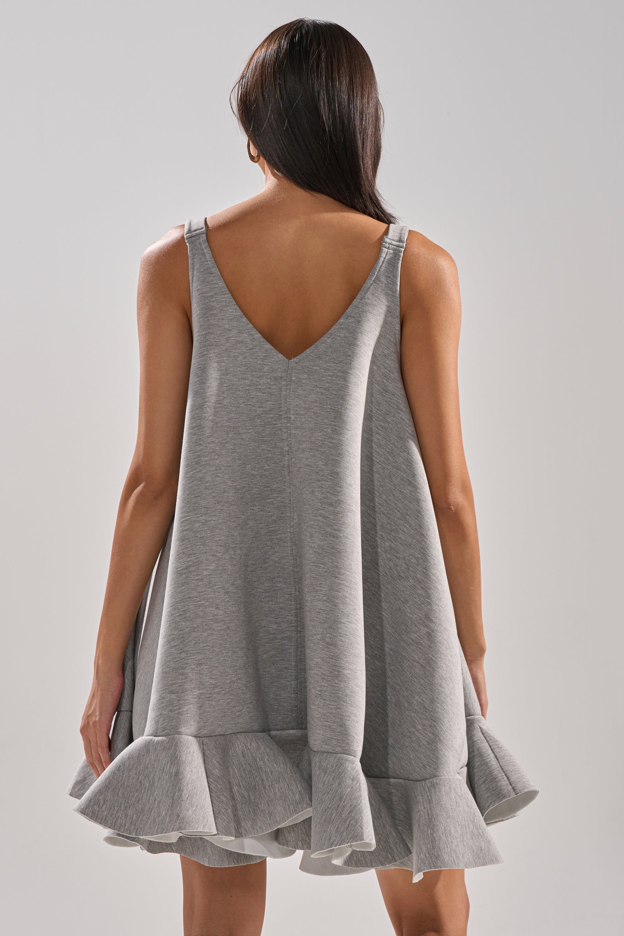 A woman stands with her back to the camera against a plain light background, wearing the IN TOO DEEP SCUBA MINI DRESS—an oversized, sleeveless grey mini dress with a deep V-back and ruffled hem.