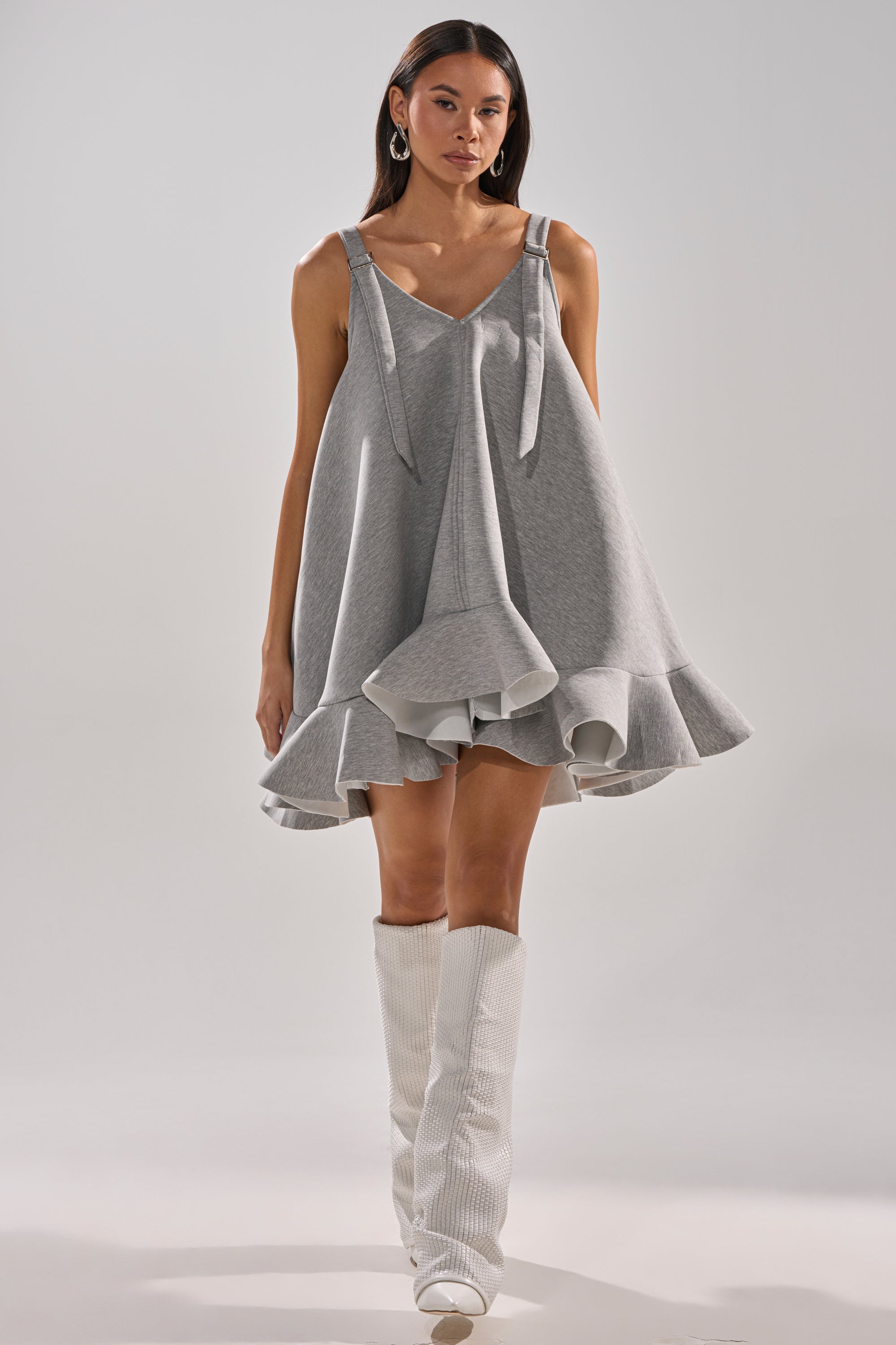A woman stands confidently in the IN TOO DEEP SCUBA MINI DRESS with bow straps and ruffled hem, styled with tall white textured boots and hoop earrings against a plain light background.