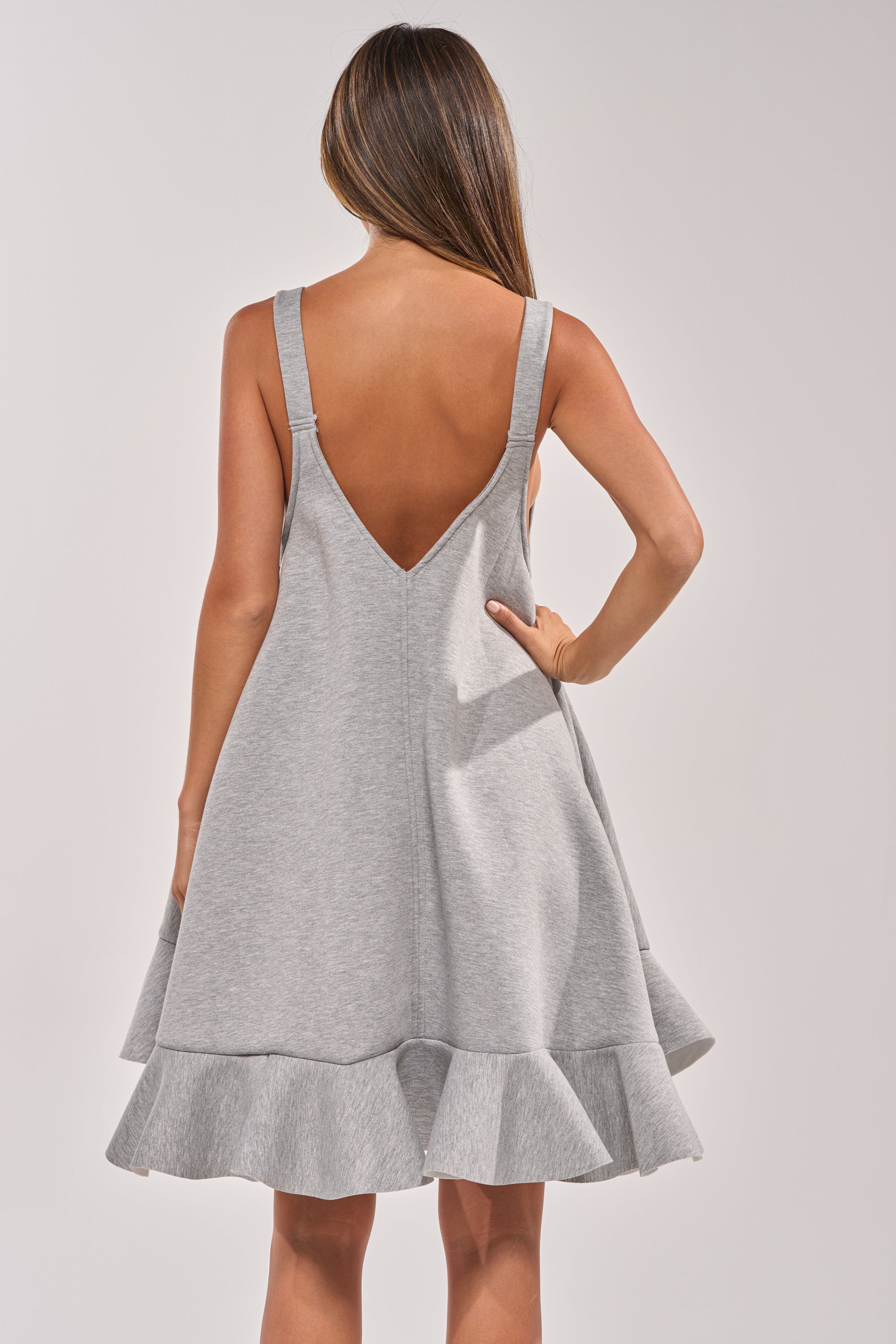 A woman with long brown hair models the IN TOO DEEP SCUBA MINI DRESS in grey, featuring a flared hem and ruffle detail. She poses with one hand on her hip, facing away for a chic, casual everyday style. 5