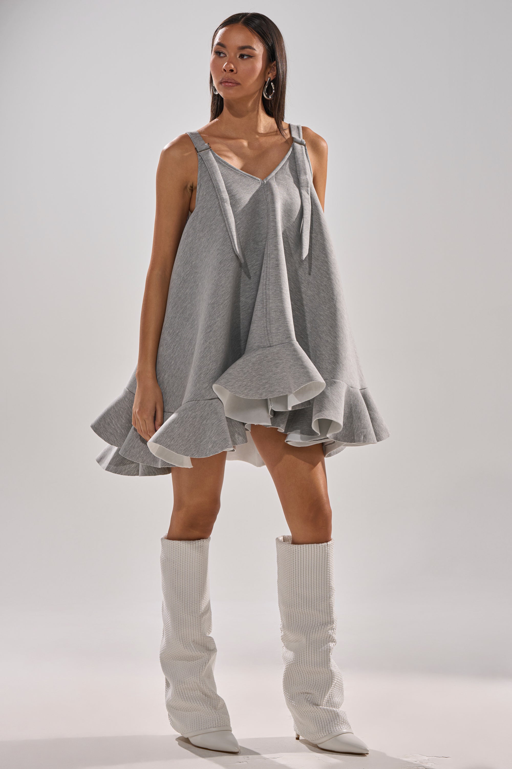 A woman models the IN TOO DEEP SCUBA MINI DRESS, a loose, light gray oversized mini dress with ruffled hem, spaghetti straps, and a V-neckline, paired with tall white textured boots against a plain background.