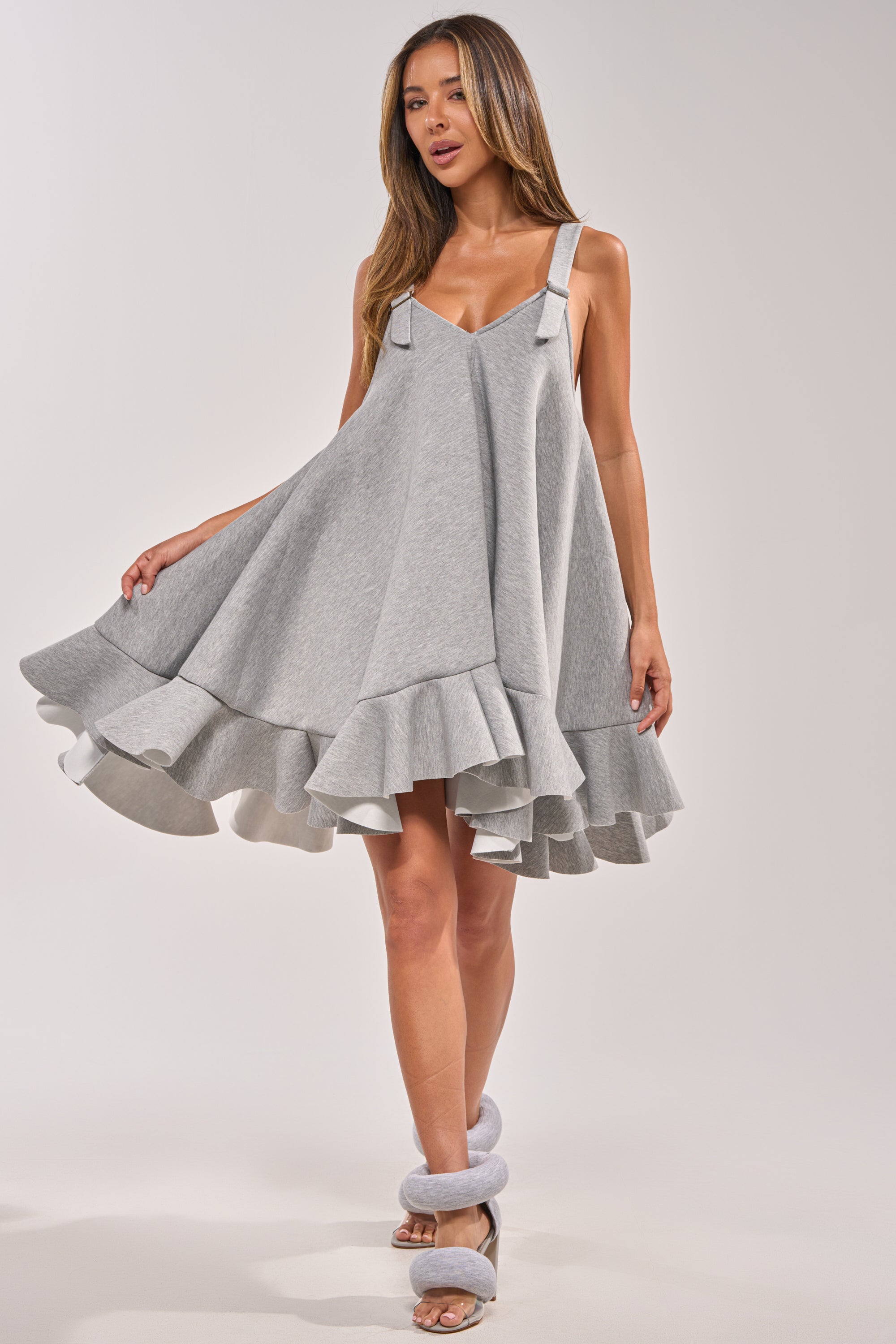 A woman poses in the IN TOO DEEP SCUBA MINI DRESS, a light gray mini dress with flared ruffled hem and bow straps, paired with matching gray open-toe heels against a plain light background. 6