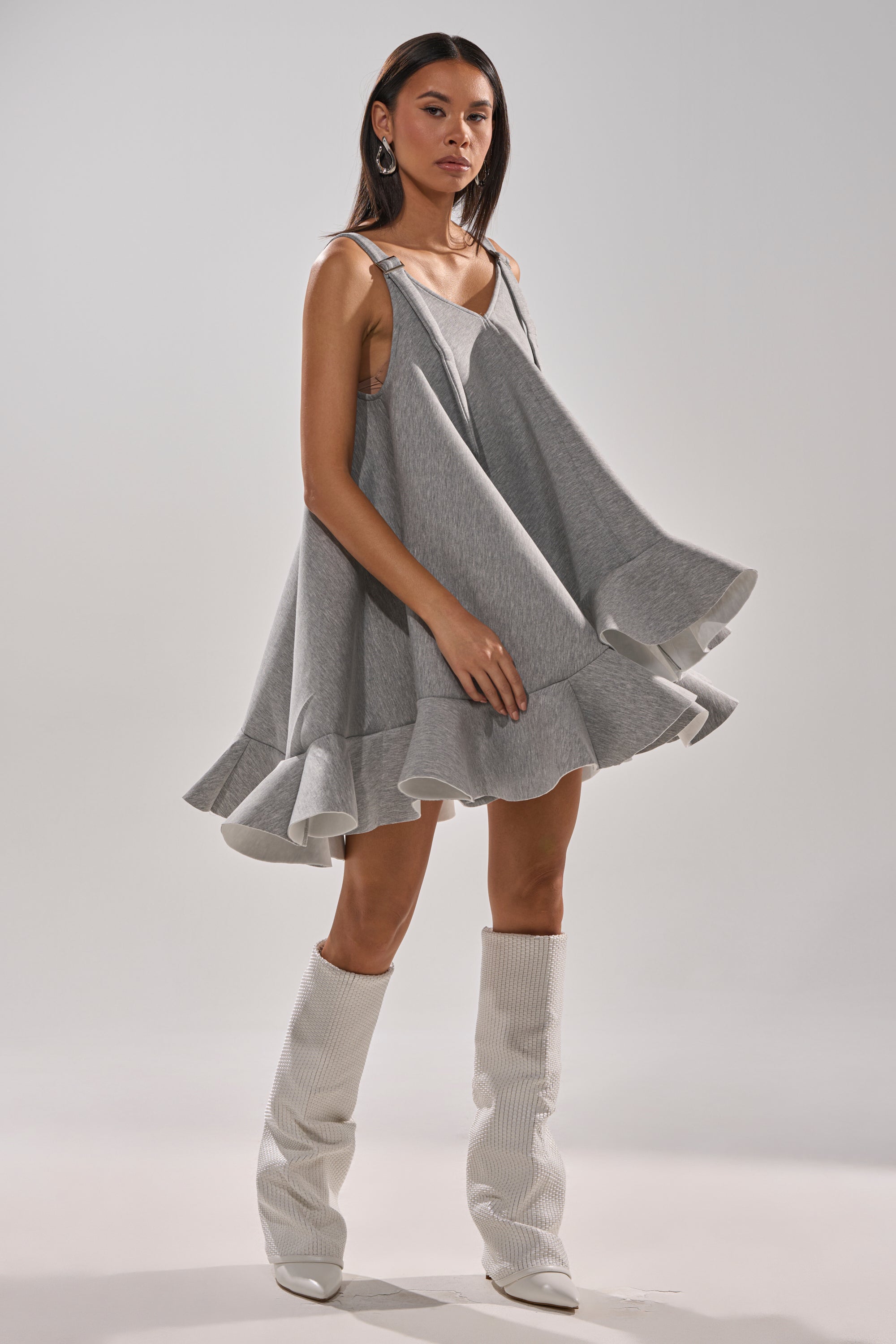 A woman models the IN TOO DEEP SCUBA MINI DRESS, a sleeveless, oversized gray mini with a ruffled hem, styled with white knee-high boots. She stands against a plain background with her hair down and wears large hoop earrings.