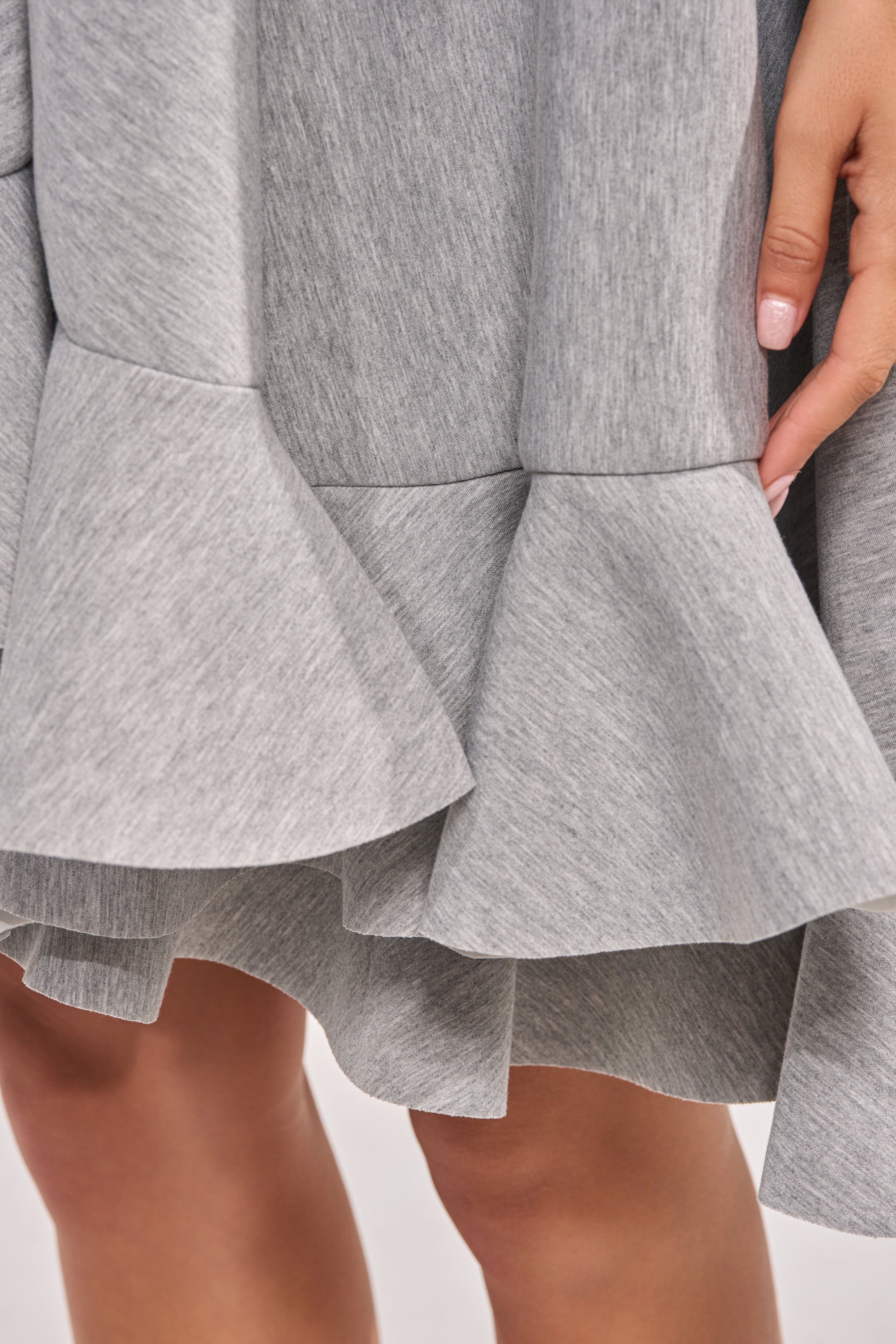A close-up of the IN TOO DEEP SCUBA MINI DRESS shows its gray a-line silhouette and layered, flared ruffles. A hand with light pink nail polish holds a ruffle. With a neutral background, it’s perfect for casual, everyday wear. 7