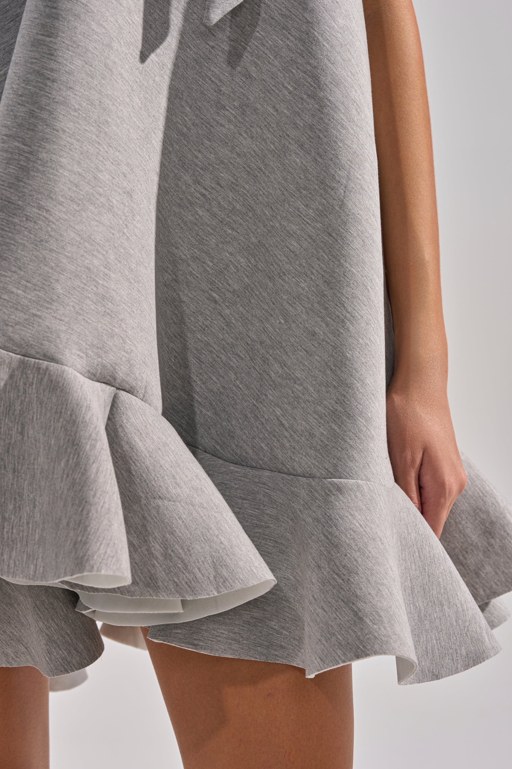 Close-up of a person wearing the IN TOO DEEP SCUBA MINI DRESS in light gray, featuring a ruffled hem and soft, slightly textured fabric. The arm is visible against a plain, light-colored background.