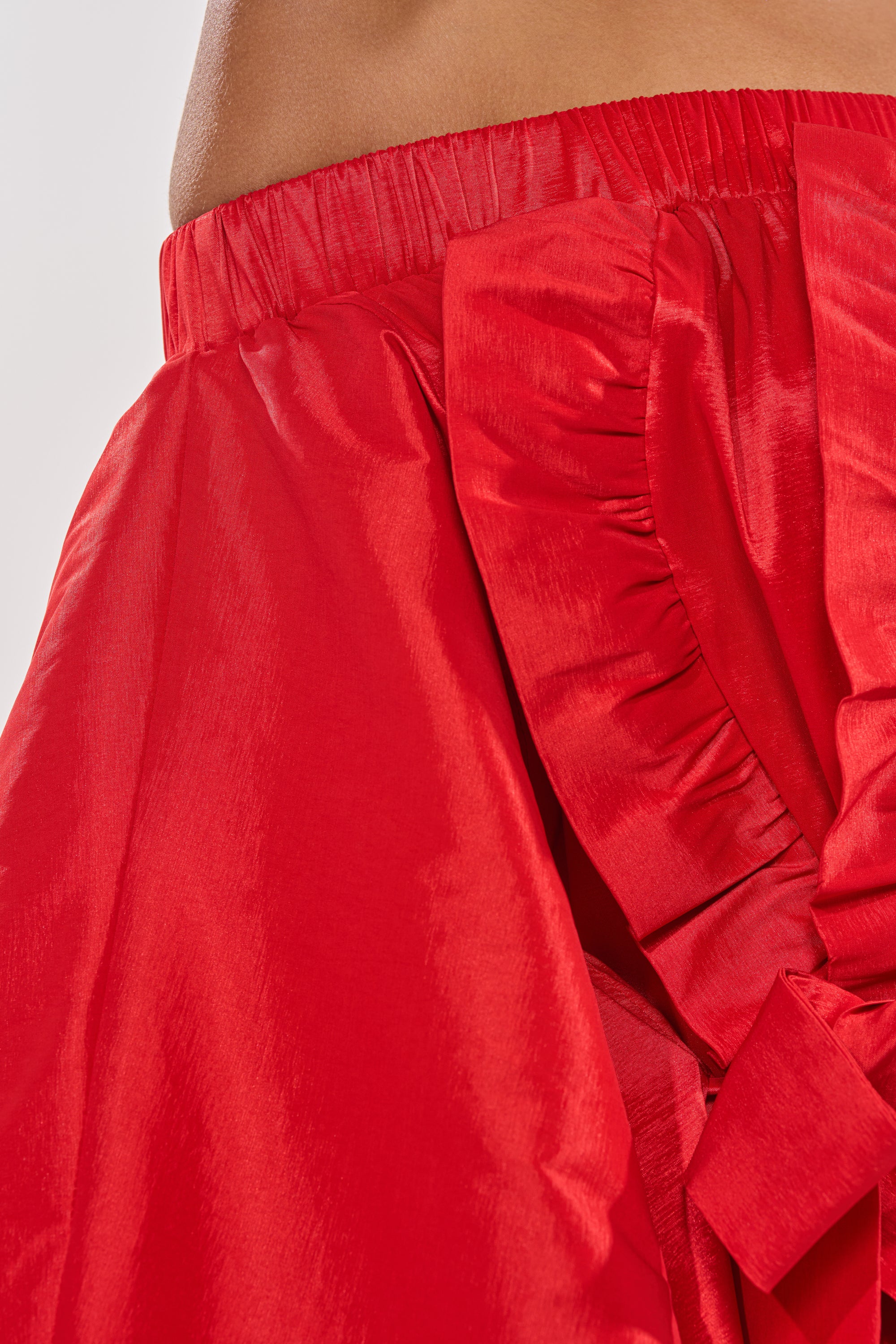 Close-up of the SHINE ON LONG SLEEVE MINI DRESS in shiny red, featuring off-the-shoulder style, ruffled and gathered details, elastic waistband, and a large decorative side bow. Wearer's skin is visible at the neckline. 9