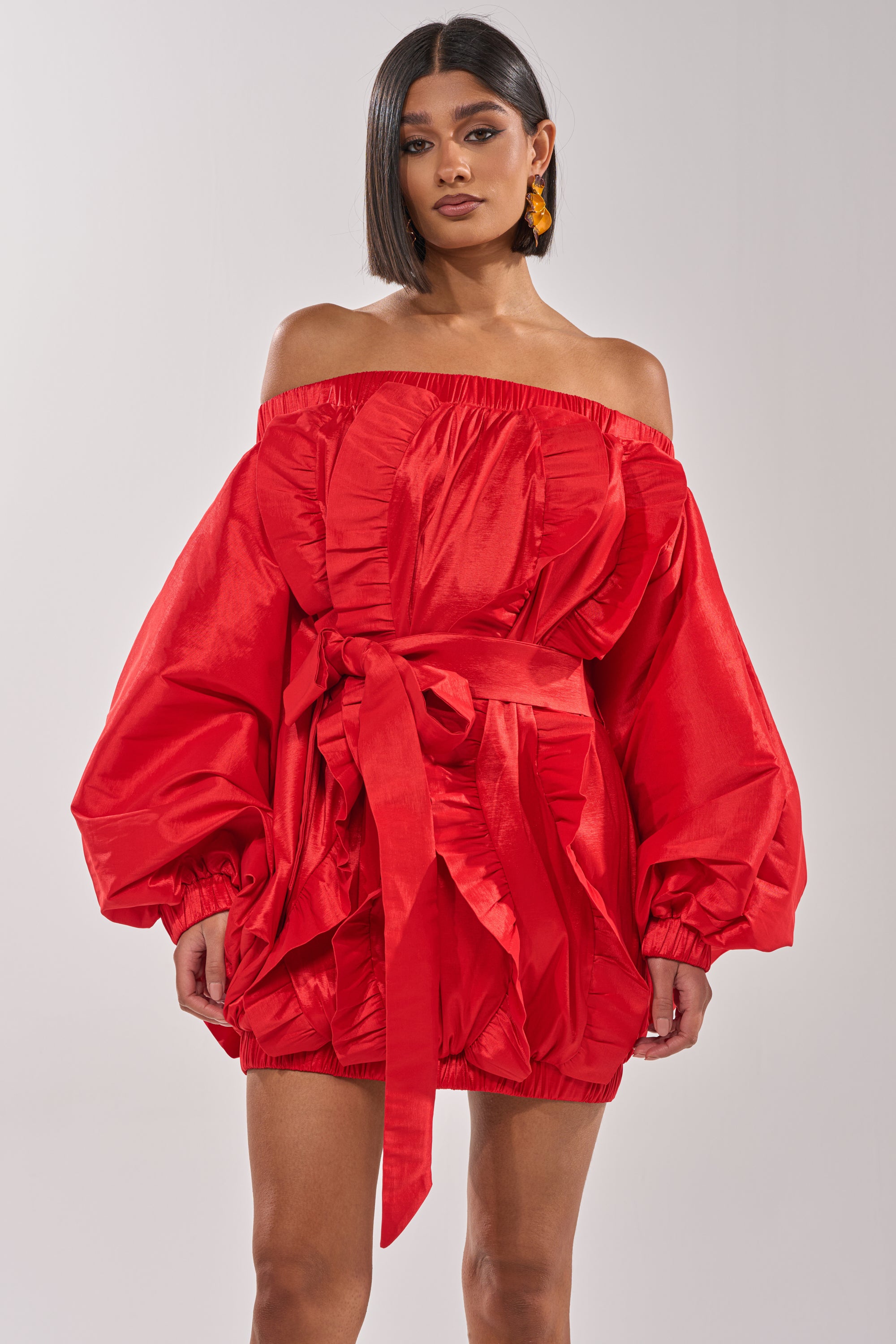 A woman models the SHINE ON LONG SLEEVE MINI DRESS—an off-the-shoulder red mini with puffy sleeves, ruffled bodice, cinched waist, and gathered hem—perfect for evening events. Her straight, shoulder-length hair adds a chic touch. 2