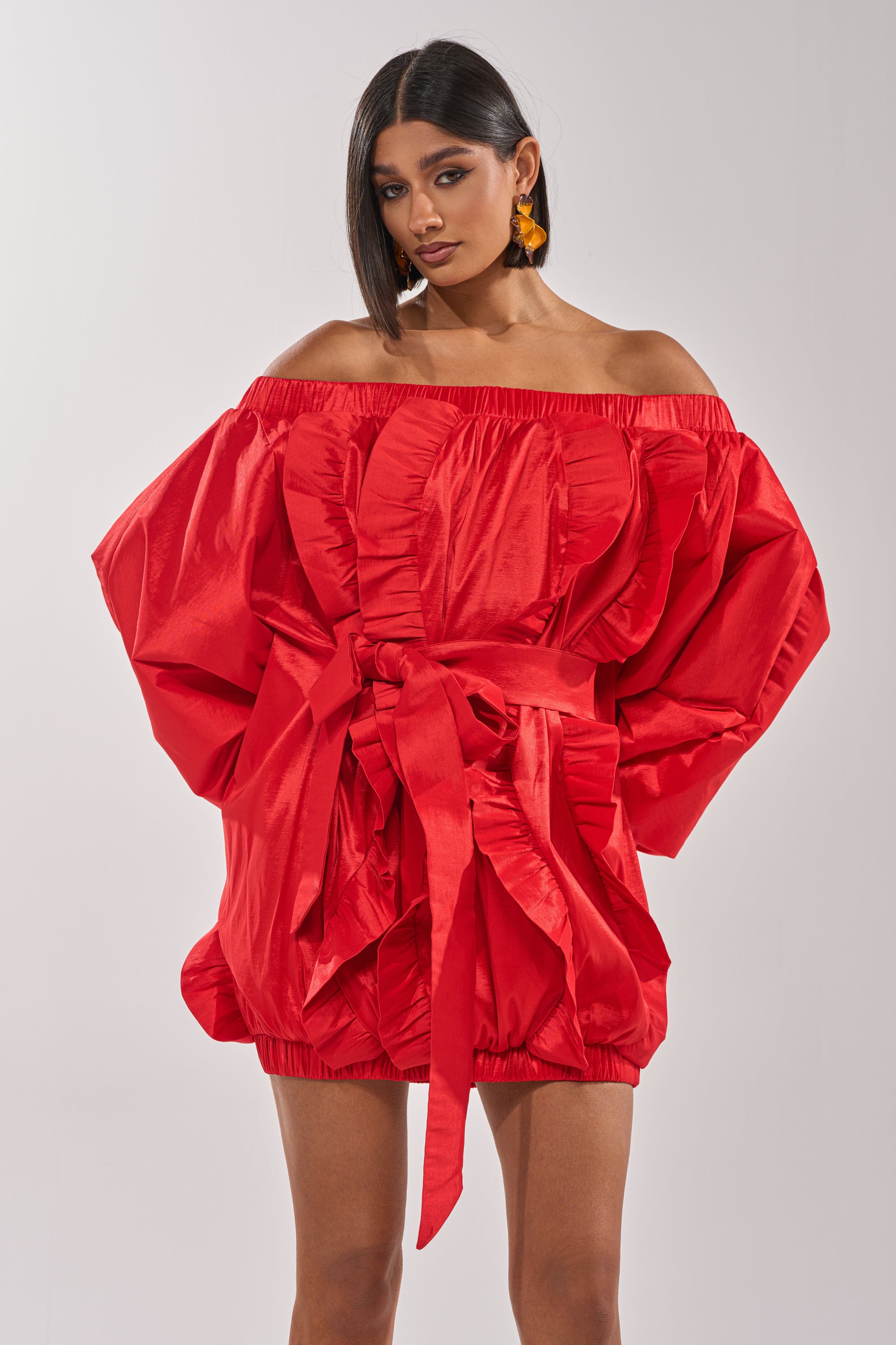 A woman models the SHINE ON LONG SLEEVE MINI DRESS, a striking red off-the-shoulder mini with voluminous sleeves and a wide sash, perfect for making an evening statement, set against a plain background. 1