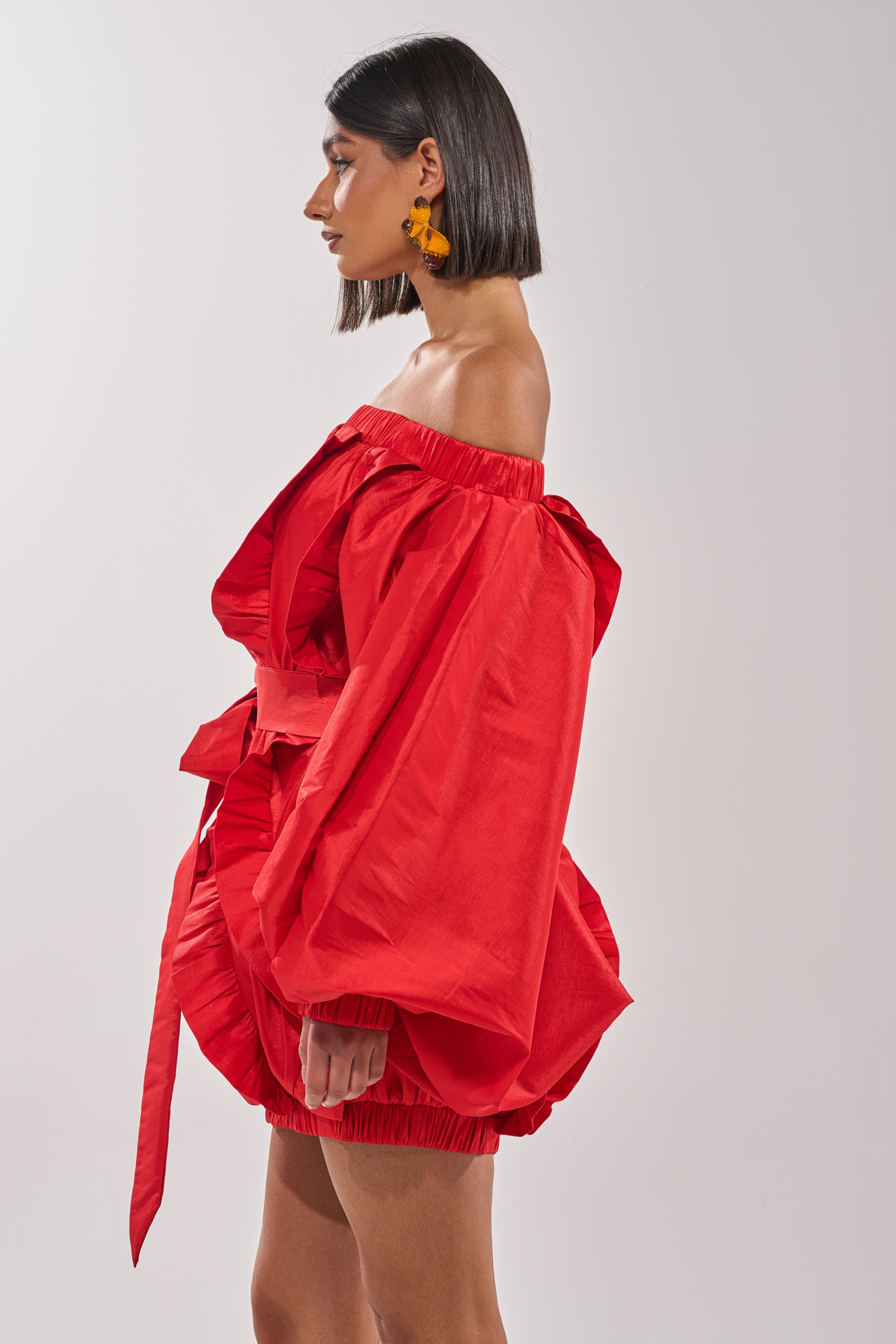 A woman with shoulder-length dark hair stands sideways against a plain light background, wearing the SHINE ON LONG SLEEVE MINI DRESS—a bright red, off-the-shoulder mini with voluminous sleeves and a belted waist, ideal for evening events. 3