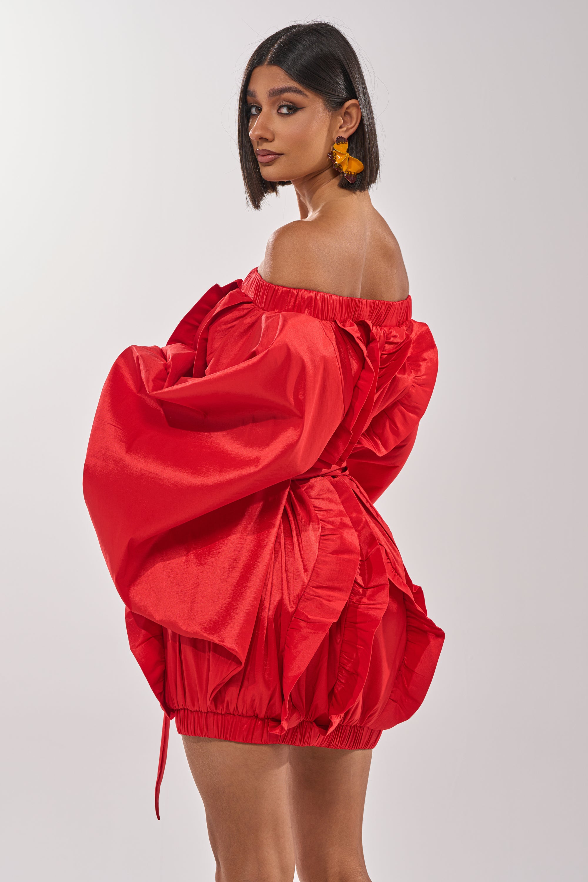 A woman with a chin-length bob models the SHINE ON LONG SLEEVE MINI DRESS—a bold red off-the-shoulder mini dress with dramatic sleeves and a ruffled skirt. She looks over her shoulder against a simple light background. 4
