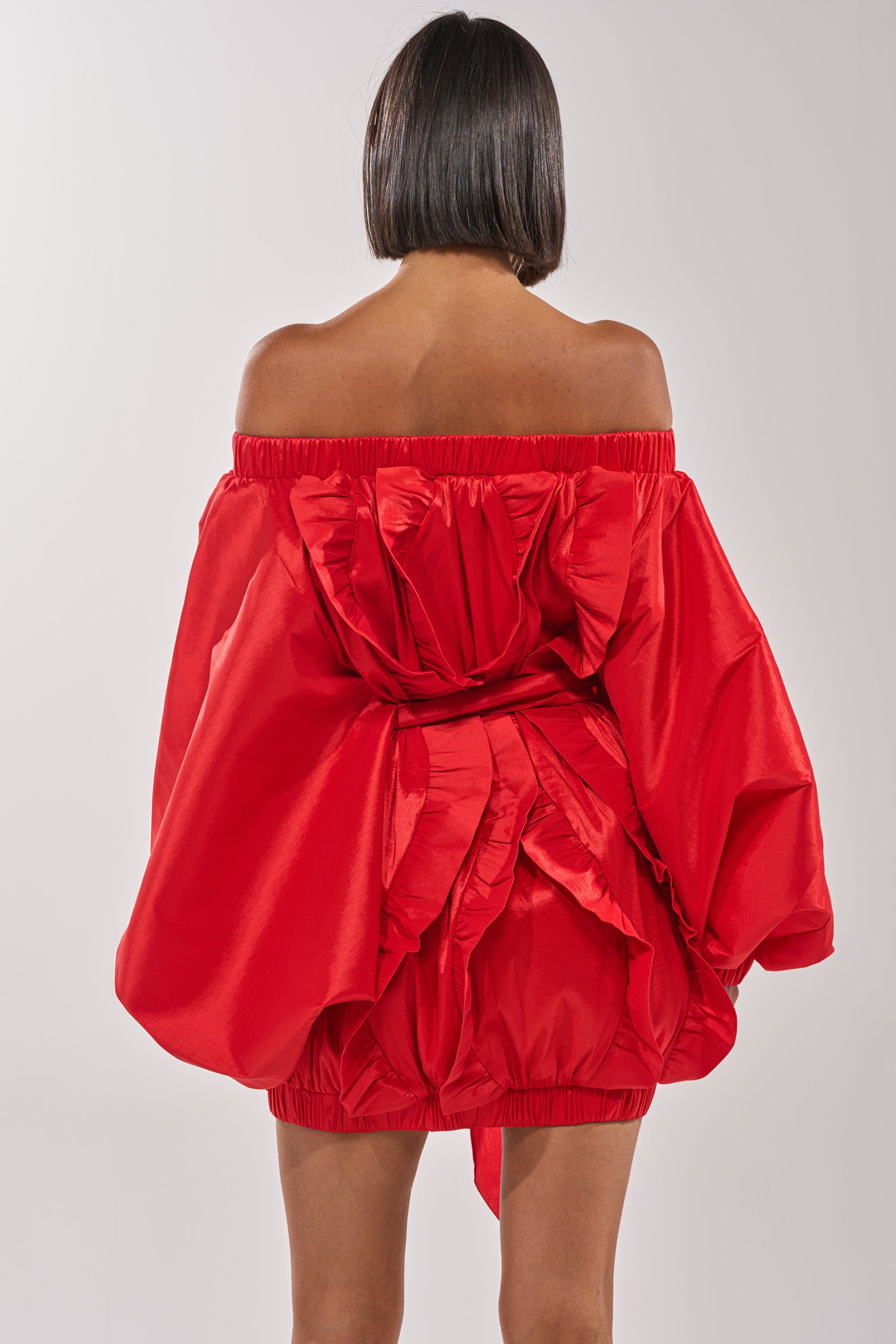 A woman with straight, chin-length dark hair faces away, wearing the SHINE ON LONG SLEEVE MINI DRESS—a bold red off-the-shoulder mini with voluminous ruffles. The plain light background highlights this striking evening statement piece. 5