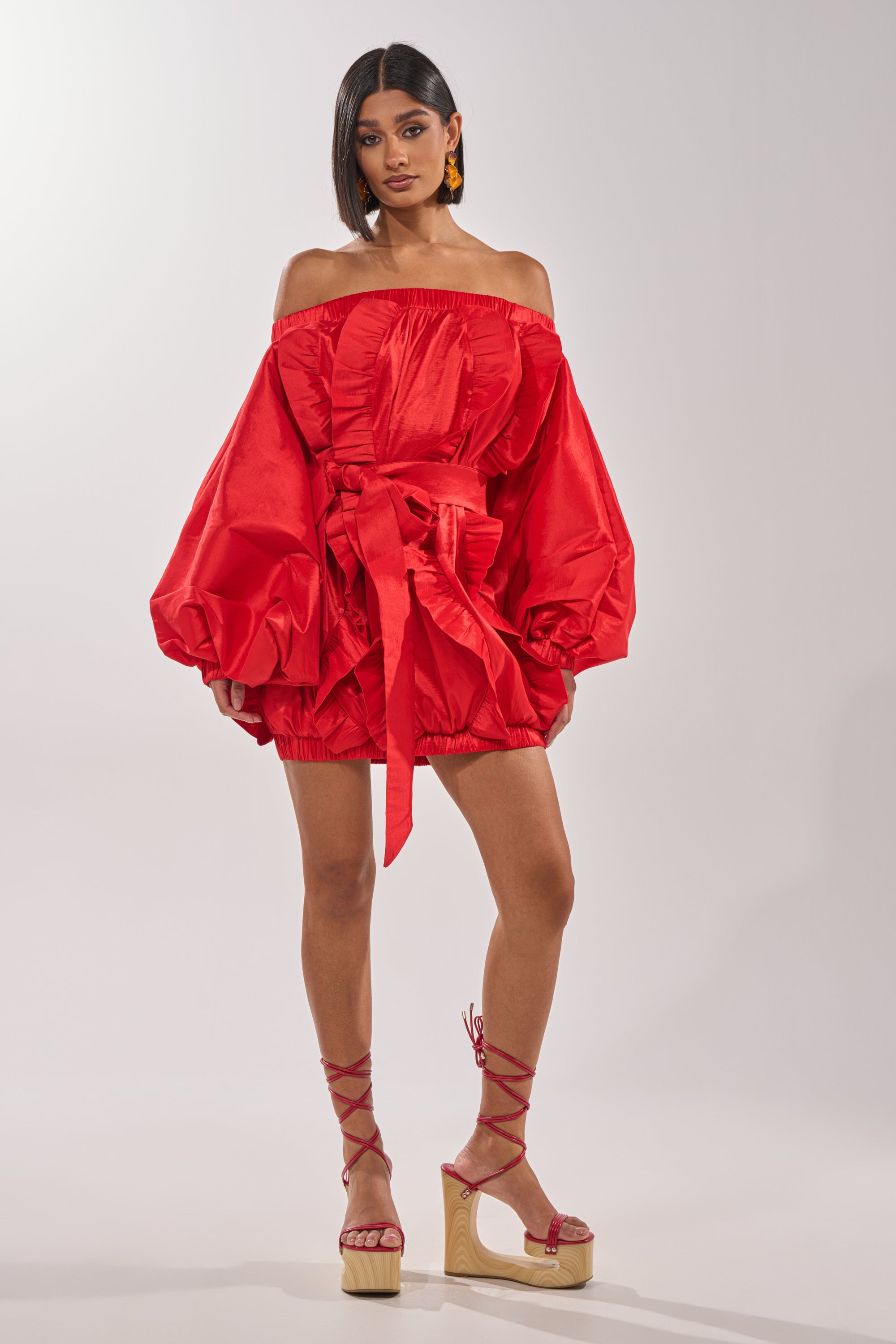 A woman wears the SHINE ON LONG SLEEVE MINI DRESS, a bold red off-the-shoulder mini with dramatic puffed sleeves and a cinched waist, paired with red lace-up platform sandals, straight dark hair, and orange earrings—a true evening statement. 7