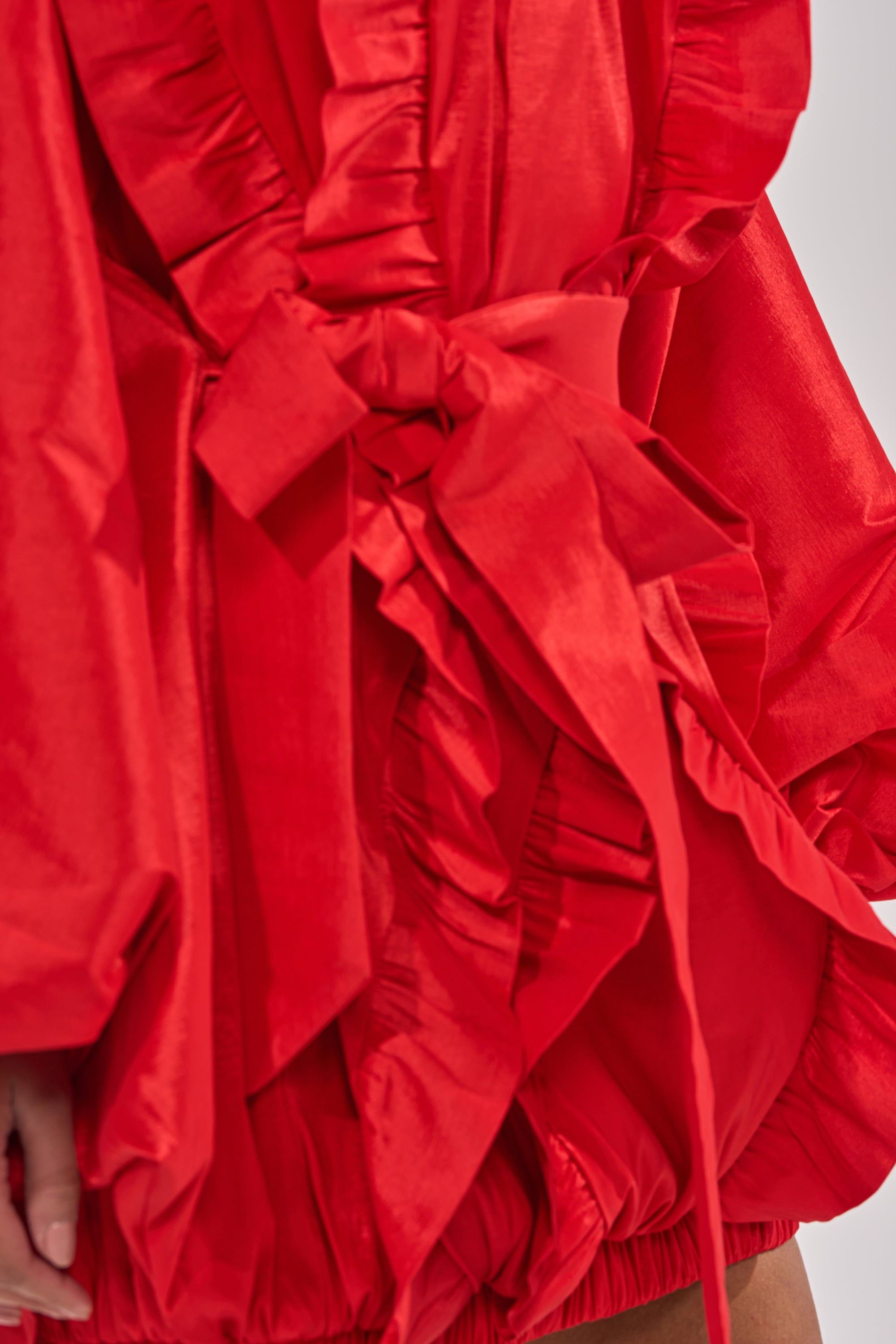Close-up of the SHINE ON LONG SLEEVE MINI DRESS in shiny red satin, featuring ruffled, gathered fabric and a large tied waist bow. The voluminous mini dress reveals part of an arm and leg for a bold statement look. 8