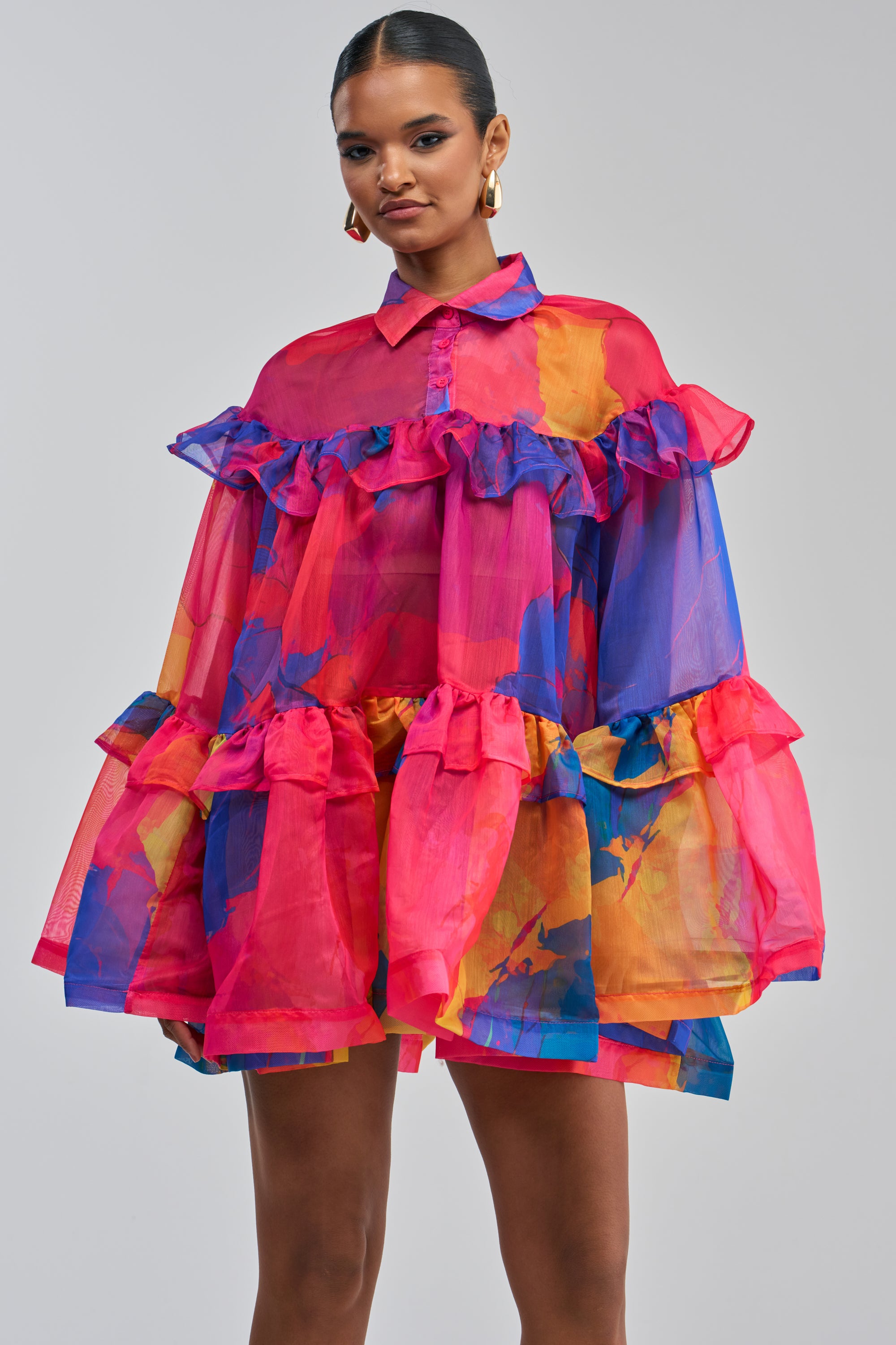 A woman stands against a plain light background wearing the CALL ME BABY CHIFFON MINI DRESS IN PINK MULTI—a sheer, colorful ruffle mini dress featuring bold pink, blue, and orange abstract patterns and a buttoned collar—styled with gold hoop earrings. This eye-catching abstract print dress makes a striking statement with its vibrant hues and playful design.