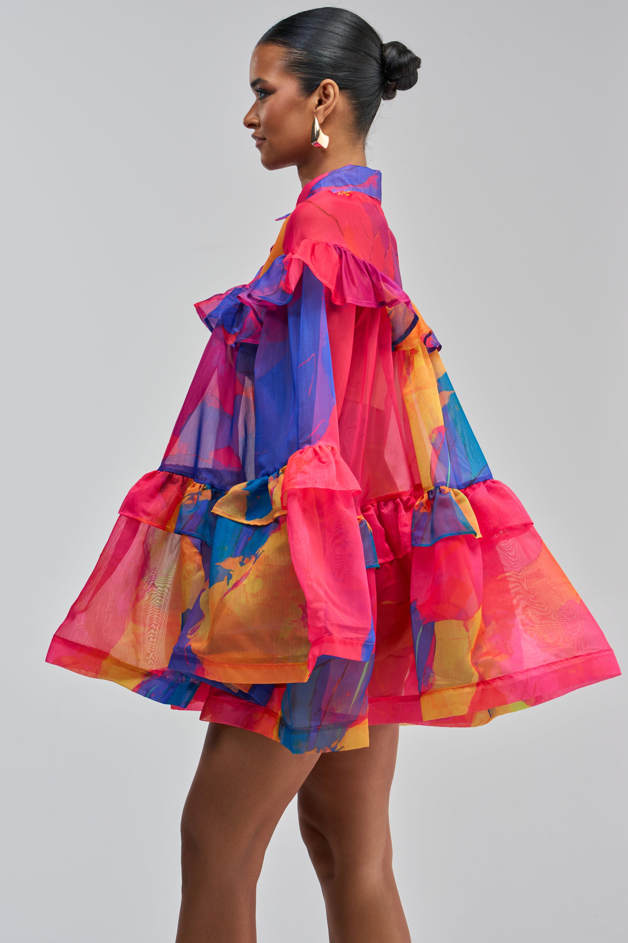 A woman stands sideways against a plain gray background, wearing the CALL ME BABY CHIFFON MINI DRESS IN PINK MULTI—a flowy chiffon mini dress featuring bold pink, blue, orange, and yellow abstract patterns. She has her hair styled in a bun and accessorizes with geometric earrings.