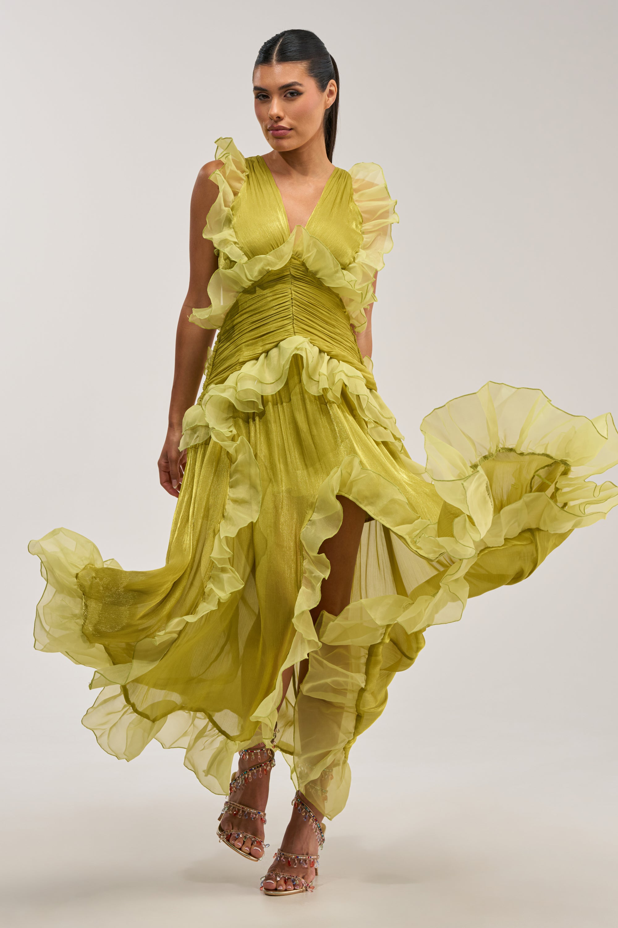 A model showcases the BEAUTIFUL GIRL FLOCKED CHIFFON MAXI DRESS IN GREEN, featuring a flowing chartreuse green silhouette with dramatic ruffled details, a deep V-neckline, ruched bodice, and billowing skirt—an elegant option for wedding guests. The look is styled with embellished strappy heels against a neutral background.
