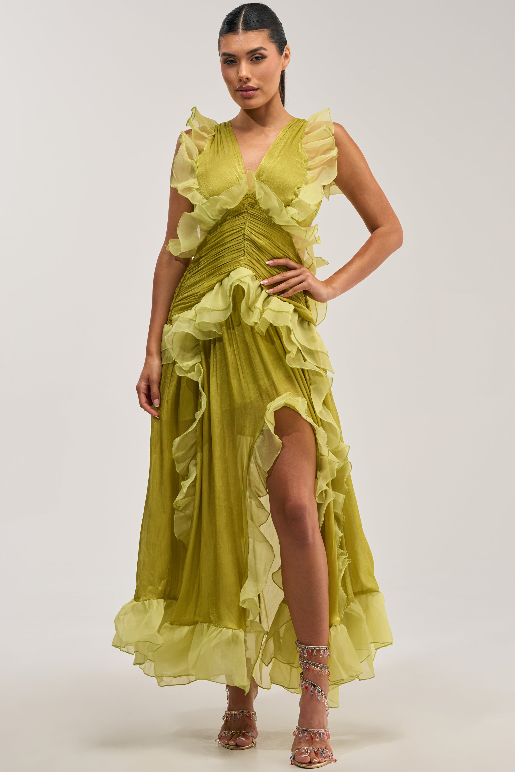 A model showcases the BEAUTIFUL GIRL FLOCKED CHIFFON MAXI DRESS IN GREEN—a vibrant chartreuse, sleeveless, floor-length chiffon gown with a pleated bodice, ruffled trim throughout, and a high front slit—paired with colorful jeweled strappy heels for an elegant formal look, all set against a plain grey background.