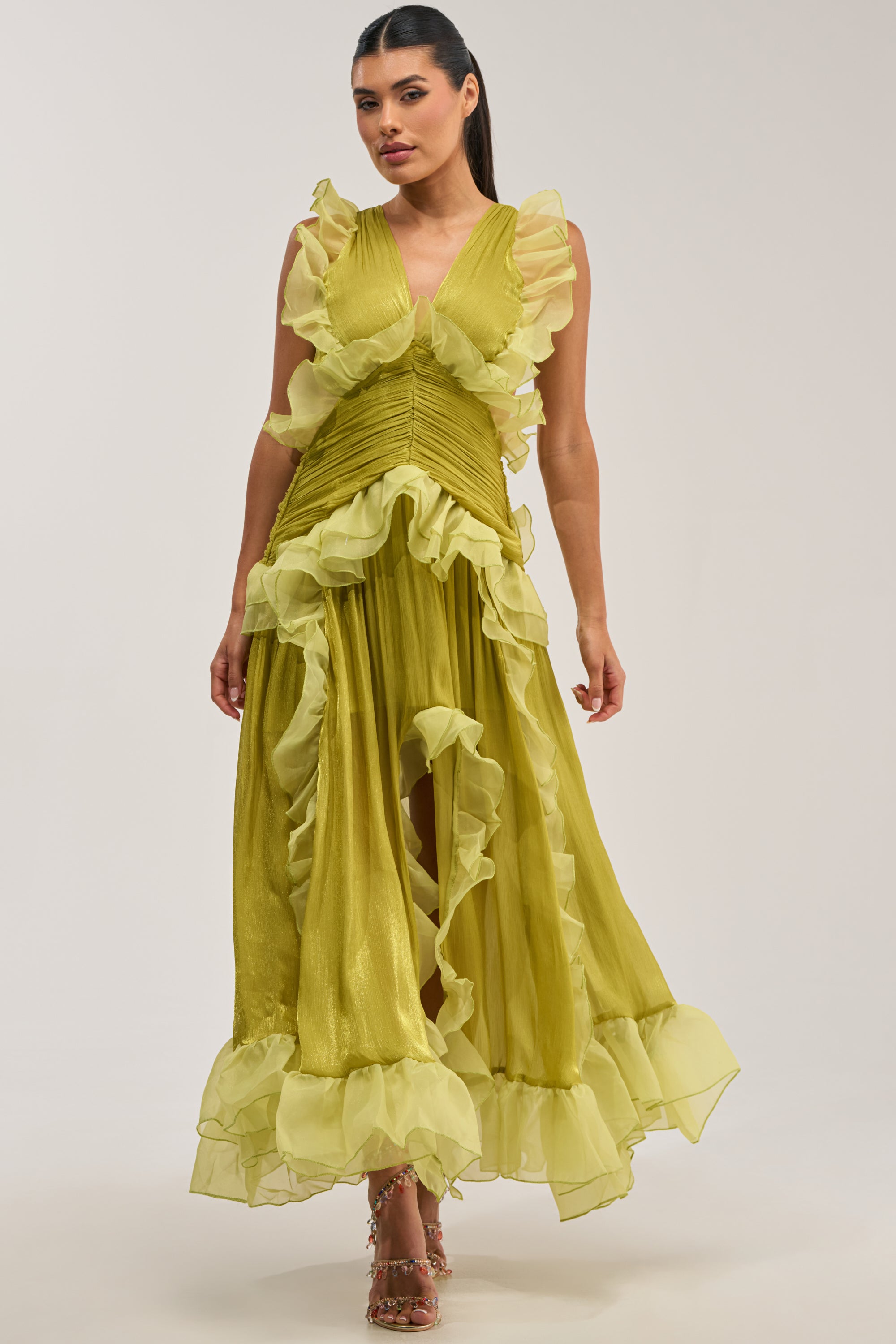 A woman wears the BEAUTIFUL GIRL FLOCKED CHIFFON MAXI DRESS IN GREEN, a sleeveless light green chiffon dress with layered, flowing fabric. Featuring a deep V-neckline and high-low hem, this elegant maxi dress is an ideal choice for a wedding guest. She stands against a plain background.