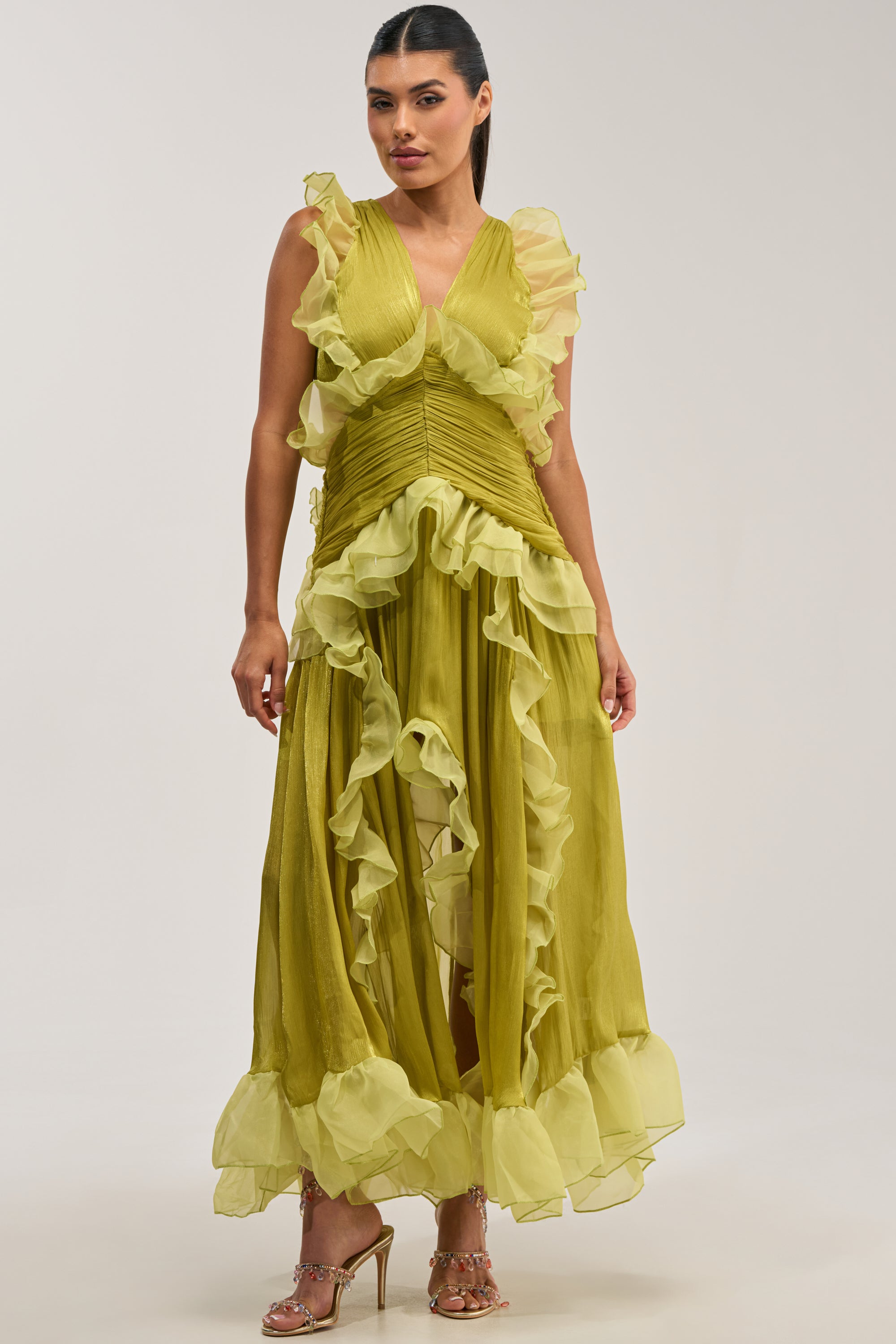 Dressed in the BEAUTIFUL GIRL FLOCKED CHIFFON MAXI DRESS IN GREEN—a sleeveless, ruffled design featuring a v-neckline and a layered, flowing skirt—the woman exudes elegance as a wedding guest. Her sleek hair is pulled back, and she stands in strappy high-heeled sandals against a plain backdrop.
