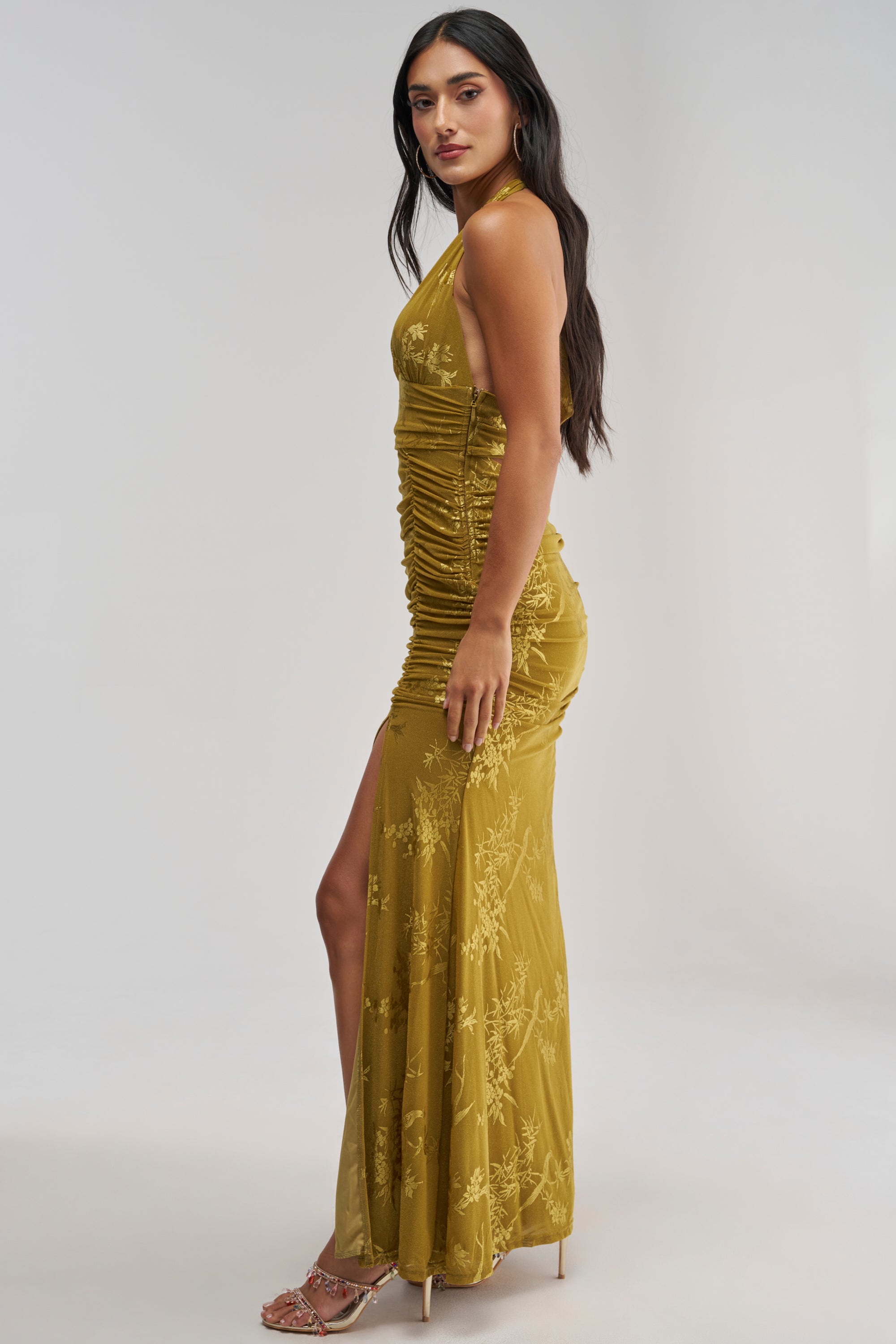 A woman stands sideways in strappy heels against a plain, light background, wearing the LIGHT LUXE MAXI DRESS IN GREEN—a sleeveless chartreuse maxi dress with a mustard yellow undertone, featuring ruched detailing and a chic side slit. Perfect as an elegant wedding guest dress.