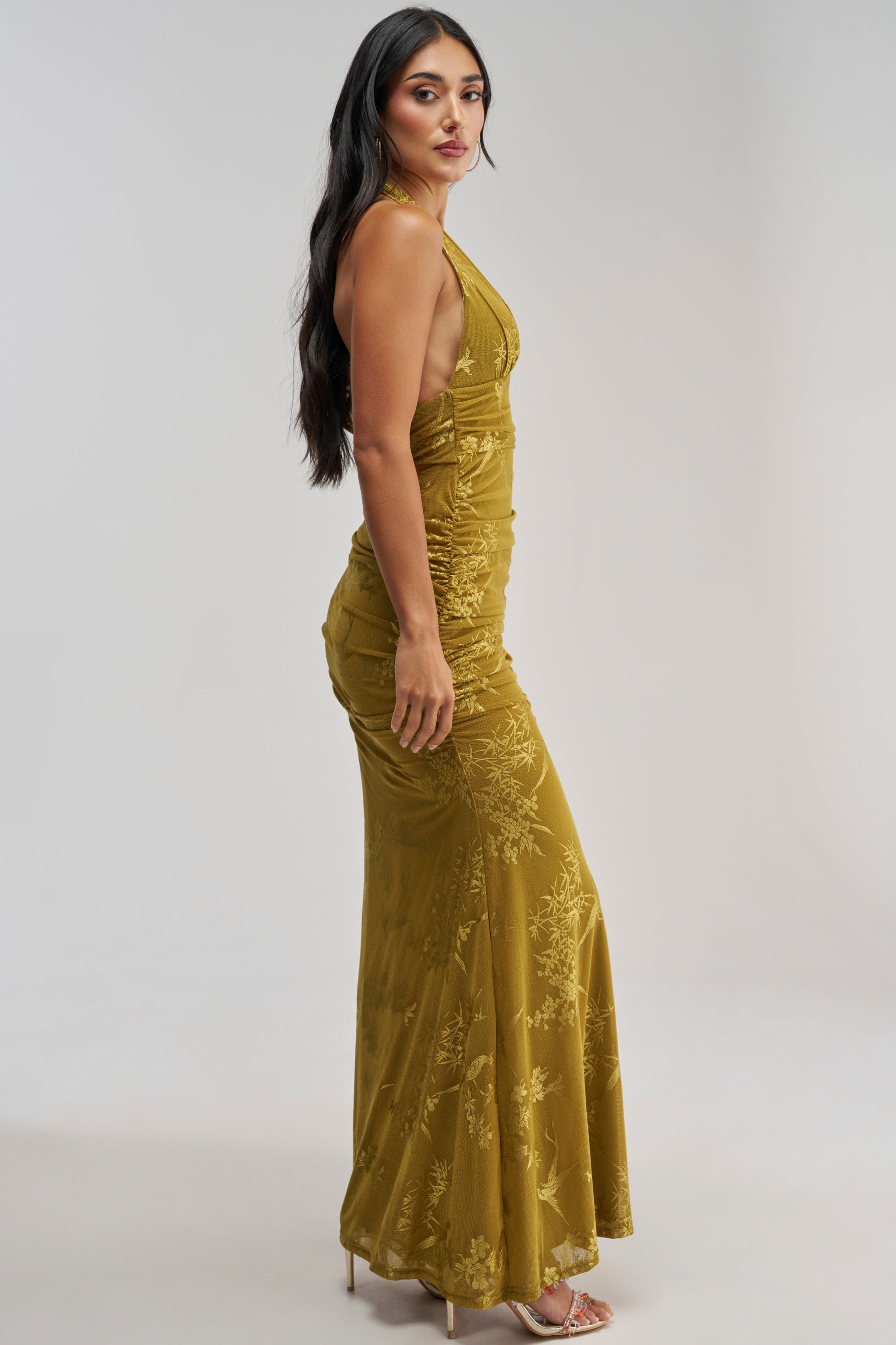 A woman poses sideways against a plain light background, wearing the LIGHT LUXE MAXI DRESS IN GREEN—an elegant, sleeveless chartreuse maxi dress featuring floral patterns and paired with high heels, making it a perfect choice for a wedding guest look.