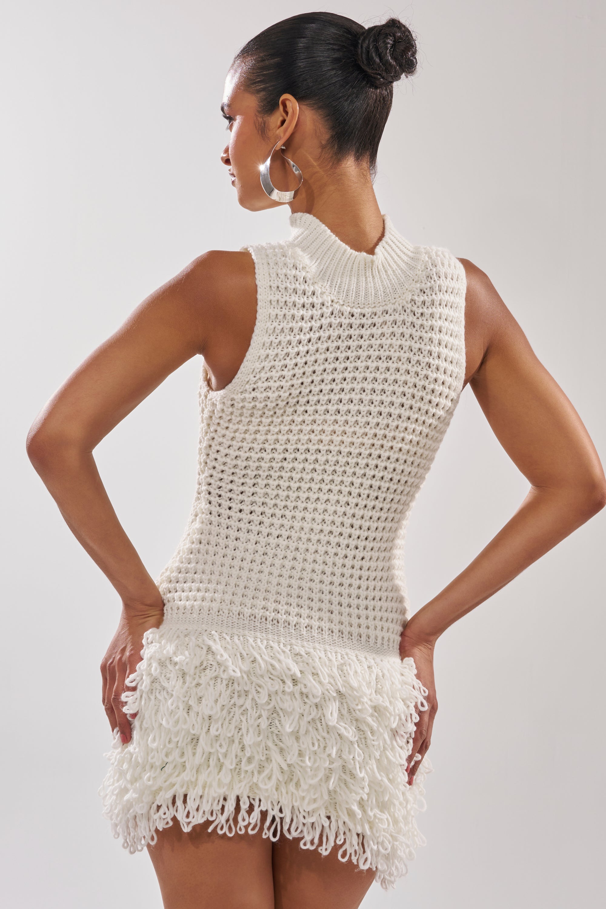 A woman with her hair in a bun models the CROCHET CUTIE MINI DRESS—an ivory knit with a textured pattern and looped fringe hem—standing hands on hips, facing away from the camera. 3