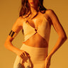 A woman with long hair poses confidently against a warm-toned background, wearing a beige cut-out dress and chain link shoulder strap accessories. She carries the TAYTAY METAL PURSE and wears a metallic arm cuff and large hoop earrings, exuding style.