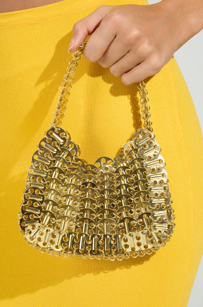 A hand holds the TAYTAY METAL PURSE, a small gold metallic bag with a chain link strap. It contrasts against a textured yellow outfit, set against a simple light background. 5